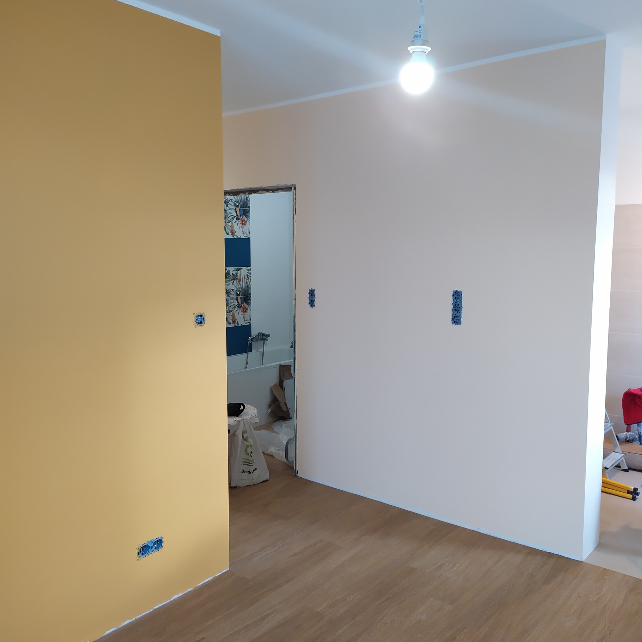 Ściana pomalowana na dwa kolory: żółty i beżowy. Widoczne gniazdka elektryczne i fragment łazienki z niebieskimi płytkami. Podłoga wyłożona panelami.