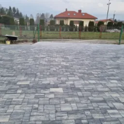 Ułożona kostka brukowa w odcieniach szarości na podwórku, z widocznym w tle domem i ogrodzeniem. W rogu taczka i wiadro po farbie.