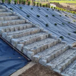 Granitowe schody na zboczu, zabezpieczone czarną folią i obsadzone roślinami. Widoczna kostka granitowa na ścieżce, w tle zieleń i sprzęt budowlany.