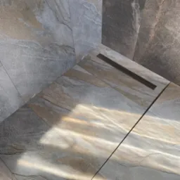 Szare i beżowe płytki imitujące kamień w kabinie prysznicowej z odpływem liniowym, oświetlone promieniami słońca.