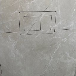 TILER PRO SERGII KUZOVINSKYI - Beżowe płytki imitujące marmur na ścianie, z zamontowanym panelem przycisku spłuczki WC. Widoczny fragment instalacji elektrycznej w górnym rogu.