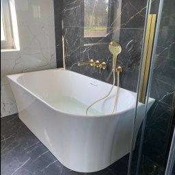 TILER PRO SERGII KUZOVINSKYI - Nowoczesna łazienka z białą wanną wypełnioną wodą, czarnymi marmurowymi płytkami i złotą armaturą, widok z boku, naturalne światło z okna.