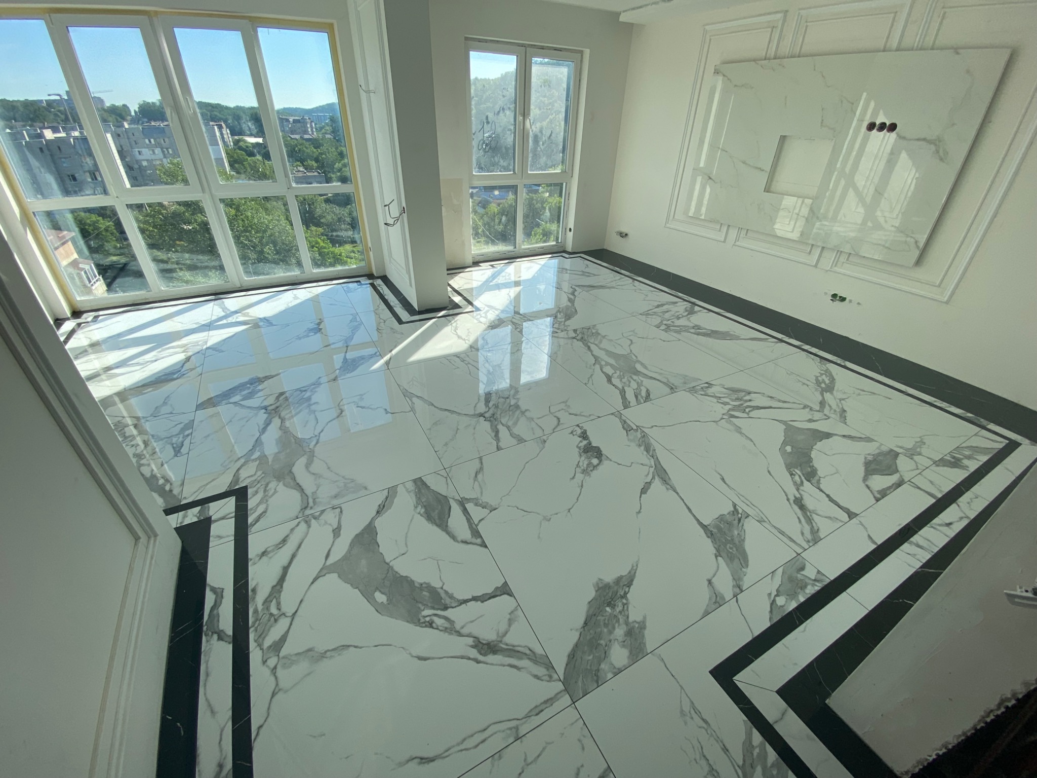 Jasna łazienka z marmurową podłogą i ścianą, duże okna z widokiem na miasto, czarne listwy przypodłogowe, przygotowane miejsce na umywalkę.