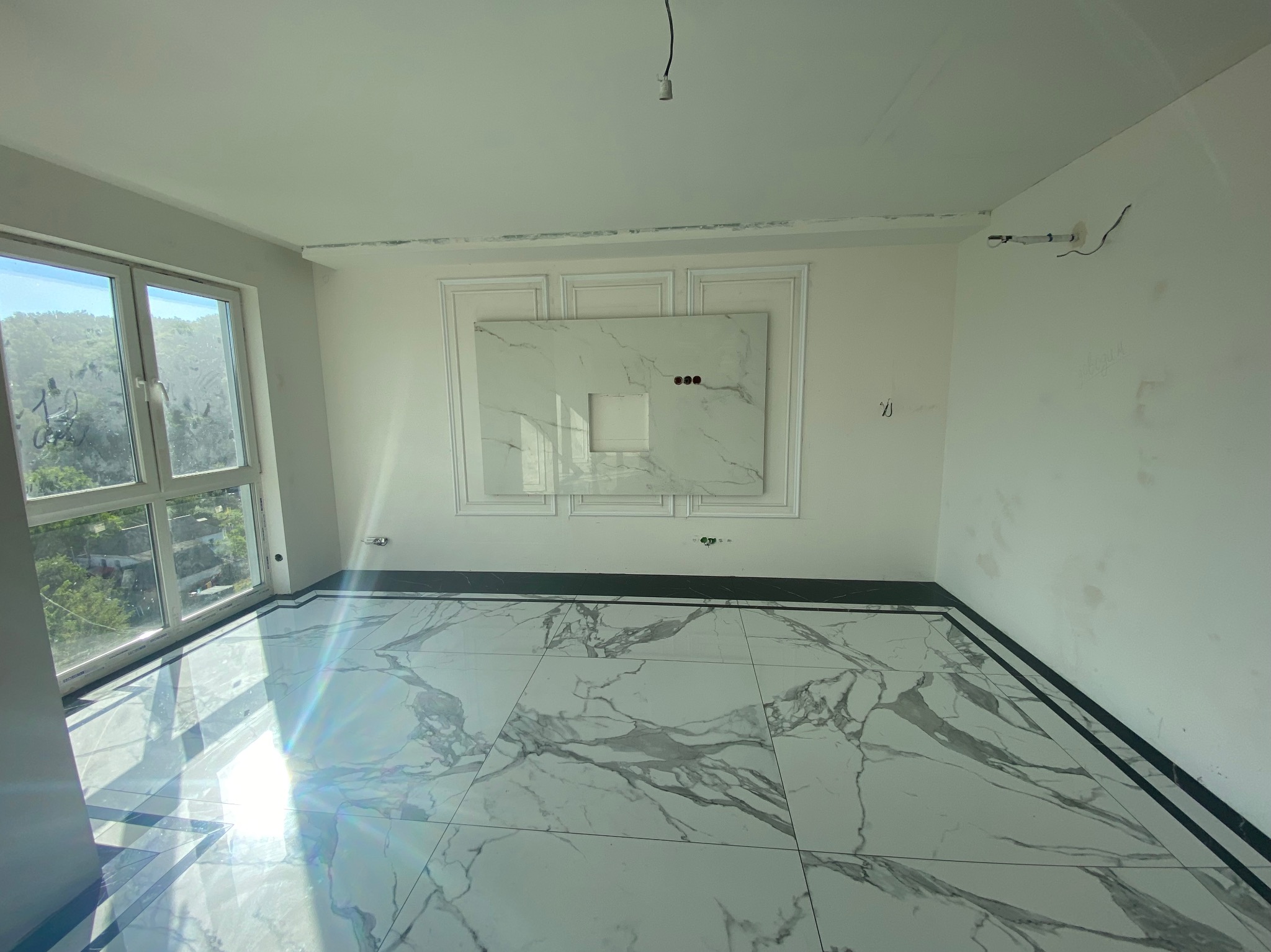 Wnętrze łazienki w trakcie wykańczania, widoczne białe płytki marmurowe na podłodze i ścianie, przygotowane pod montaż umywalki, z widocznymi przewodami elektrycznymi i oknem z widokiem na zewnątrz.