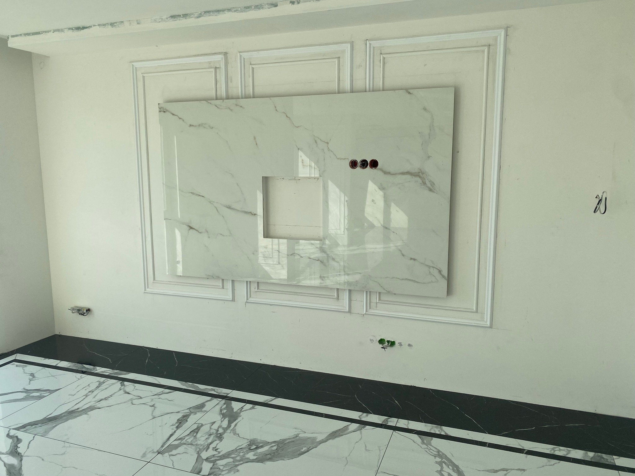 Ściana z zamontowaną płytą imitującą marmur z wycięciem na środku i trzema otworami na gniazdka elektryczne, otoczona dekoracyjnymi listwami, z podłogą wyłożoną płytkami imitującymi marmur...