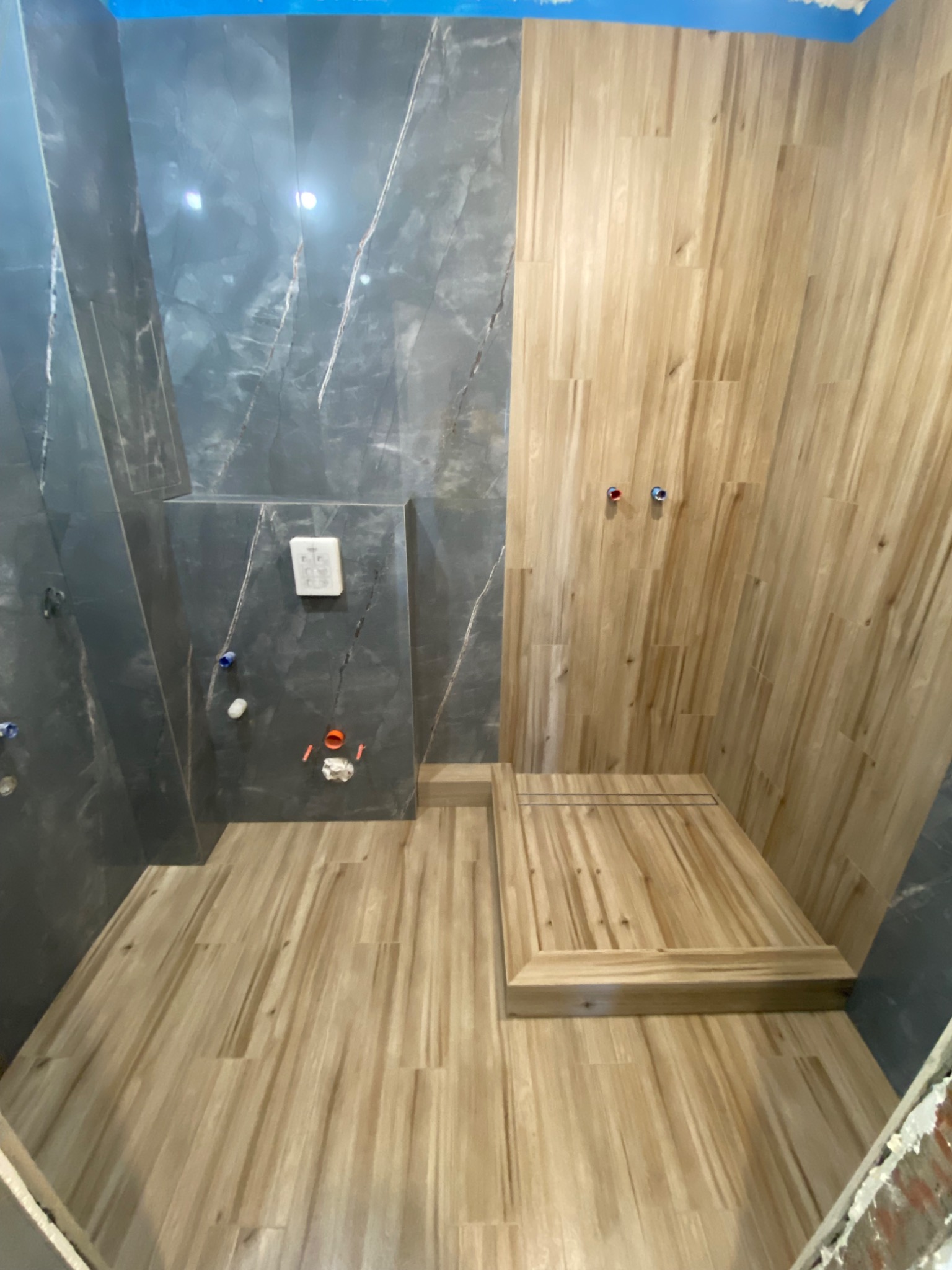 Wnętrze łazienki w Tychach w trakcie wykańczania, z drewnopodobnymi płytkami na podłodze i ścianie prysznica oraz ciemnoszarymi płytkami imitującymi marmur na pozostałych ścianach. Widoczne...