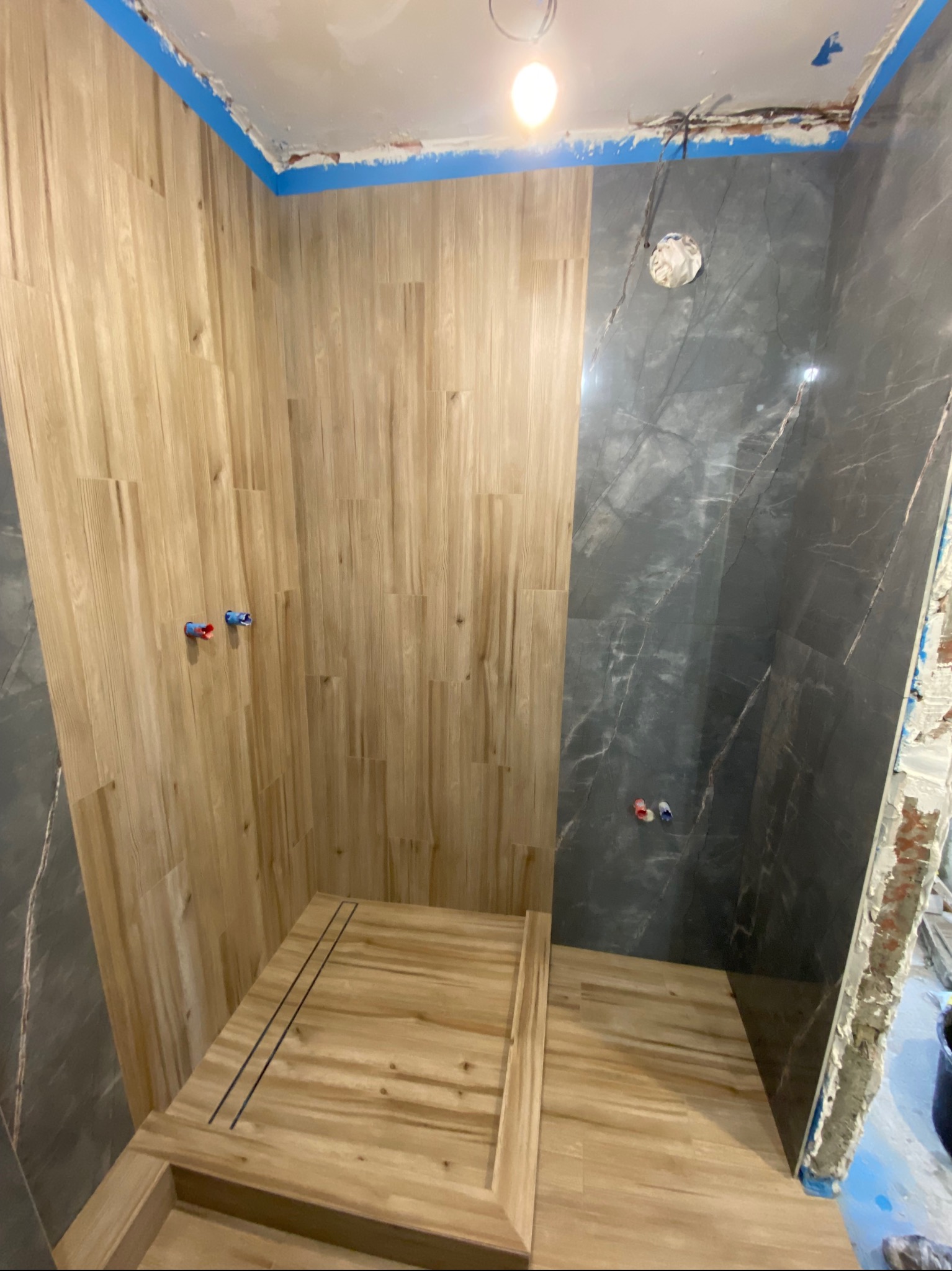 Wnętrze łazienki w trakcie prac wykończeniowych: drewnopodobne płytki na ścianach i podłodze kabiny prysznicowej z odpływem liniowym, kontrastujące z szarymi płytkami imitującymi marmur...