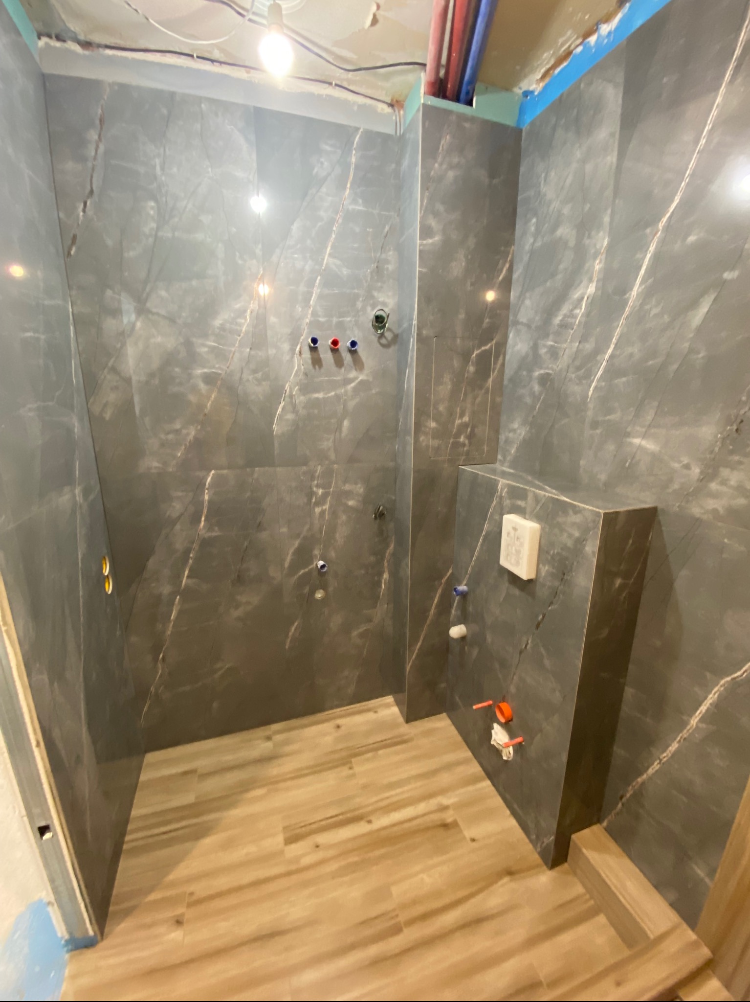 Wnętrze łazienki w trakcie remontu: szare płytki imitujące marmur na ścianach, drewnopodobne płytki na podłodze, widoczne instalacje wodne, sufit w trakcie prac wykończeniowych.
