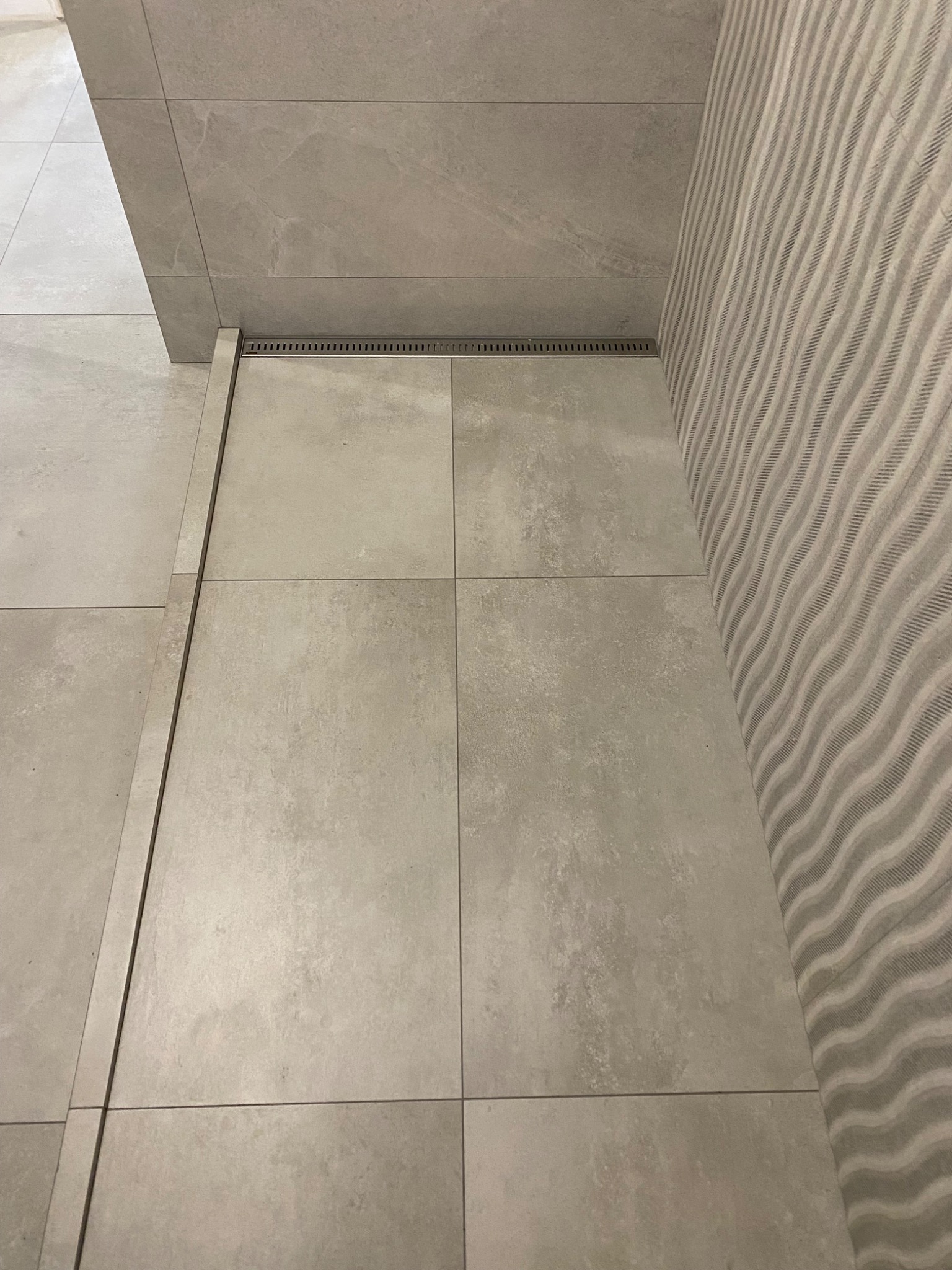 Beżowe płytki podłogowe i ścienne w łazience, z odpływem liniowym przy ścianie pokrytej tapetą w subtelne fale.