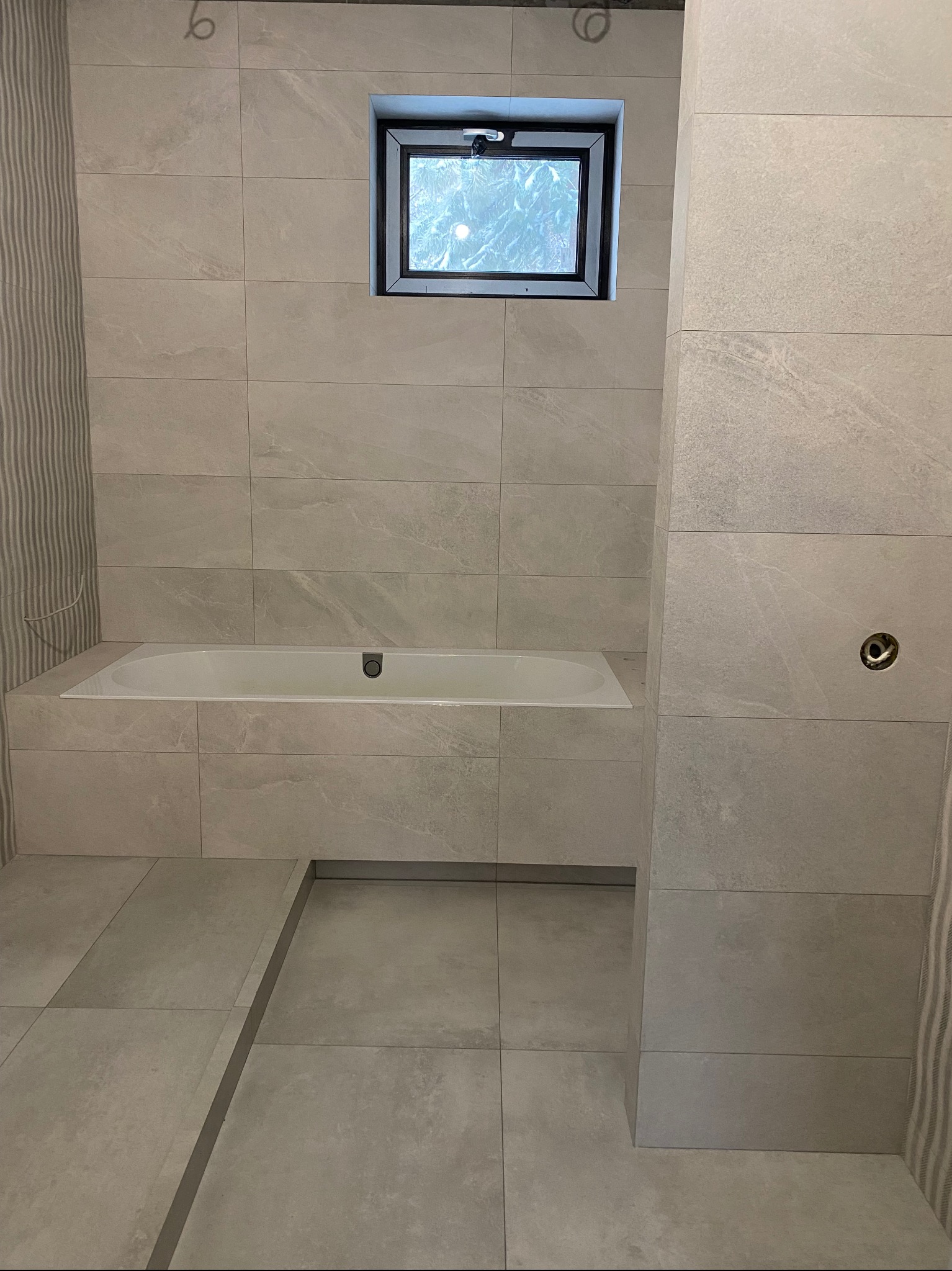 Wanna w jasnej łazience z szarymi płytkami, małe okno z czarną ramą, pionowe płytki w paski po lewej stronie, przygotowane podejście pod baterię na ścianie.