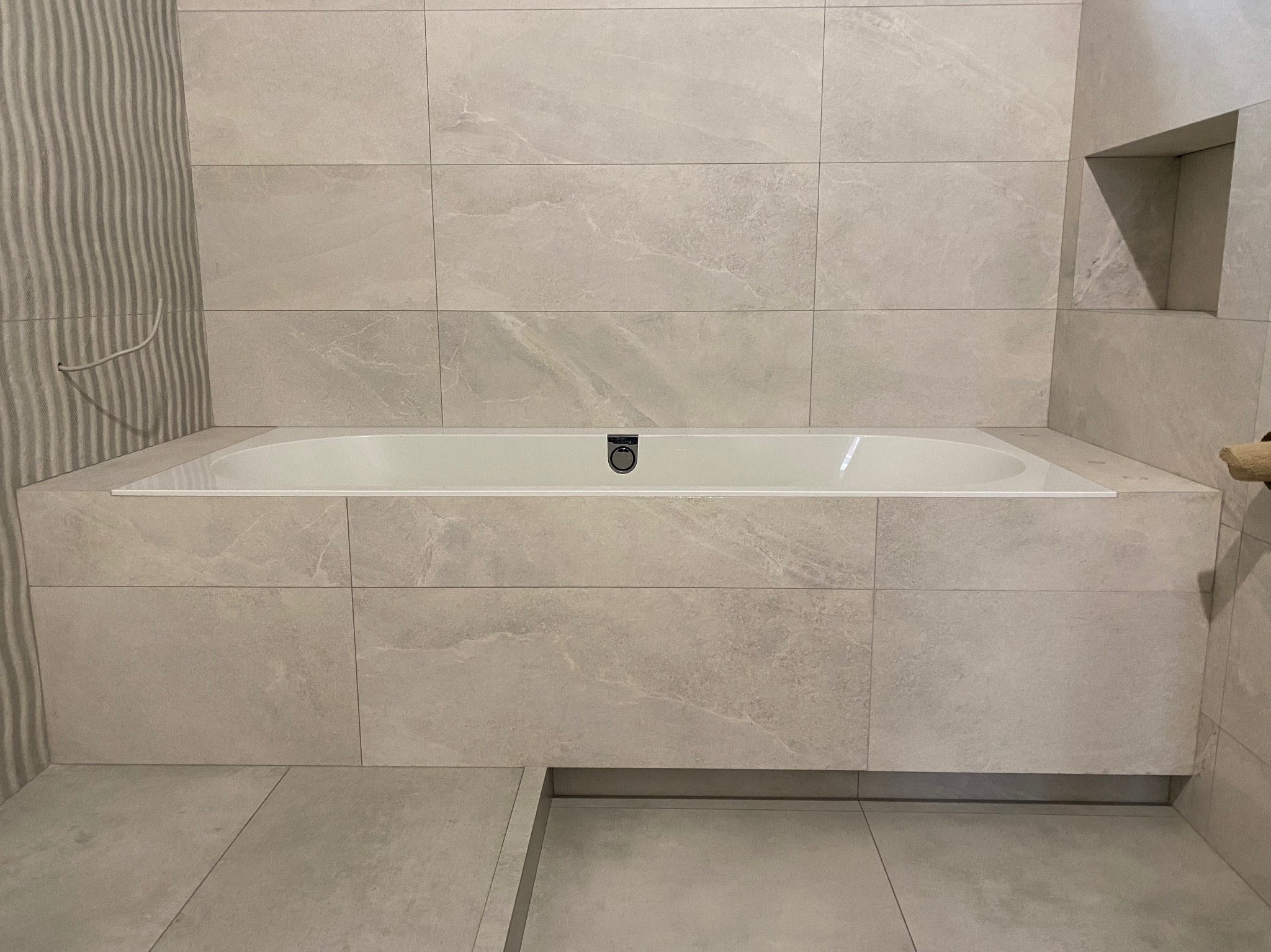 Wanna obudowana płytkami imitującymi kamień w odcieniach szarości, z ukrytym odpływem i wnęką w ścianie, minimalistyczny design.