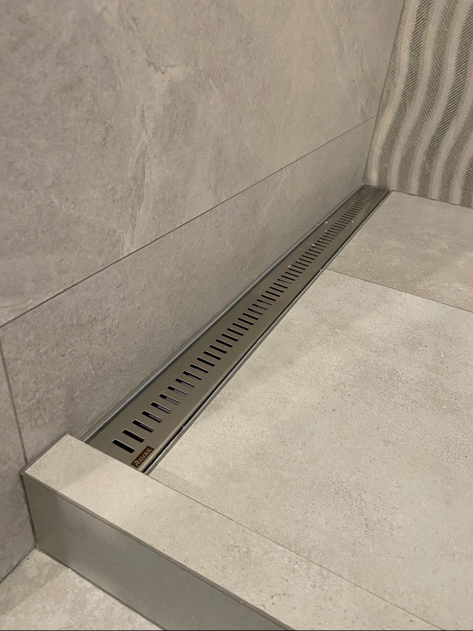 Długi, liniowy odpływ podłogowy marki RAVAK wkomponowany w szare płytki podłogowe i ścienne w łazience. Widoczny fragment ściany z dekoracyjnym wzorem.