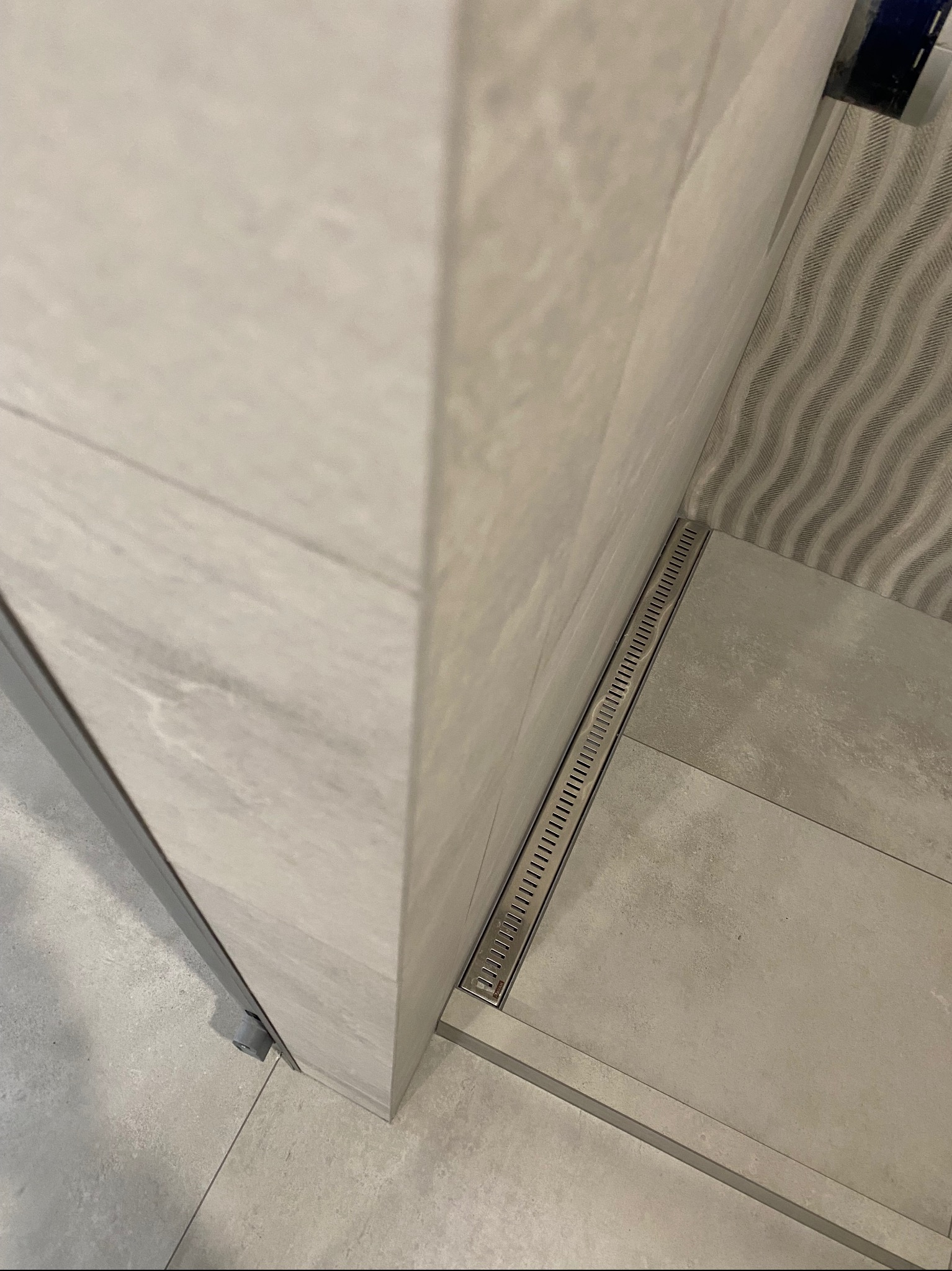 Szczegółowe ujęcie liniowego odpływu prysznicowego wkomponowanego w jasne, marmurowe płytki, z widocznym fragmentem dekoracyjnej ściany z wzorem w fale.