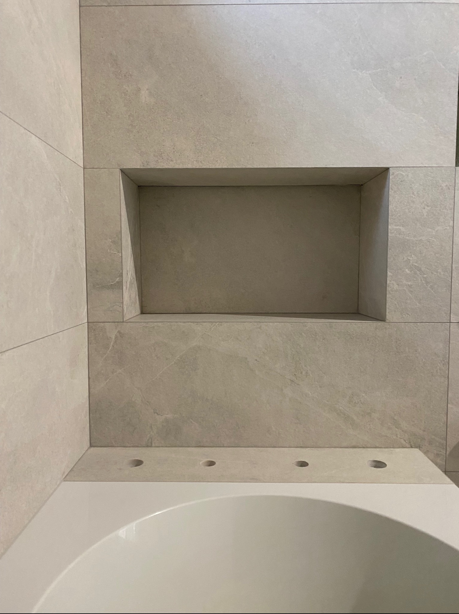 Wykończona wnęka w ścianie nad wanną, obłożona jasnymi płytkami imitującymi marmur, z widocznymi otworami na baterię.