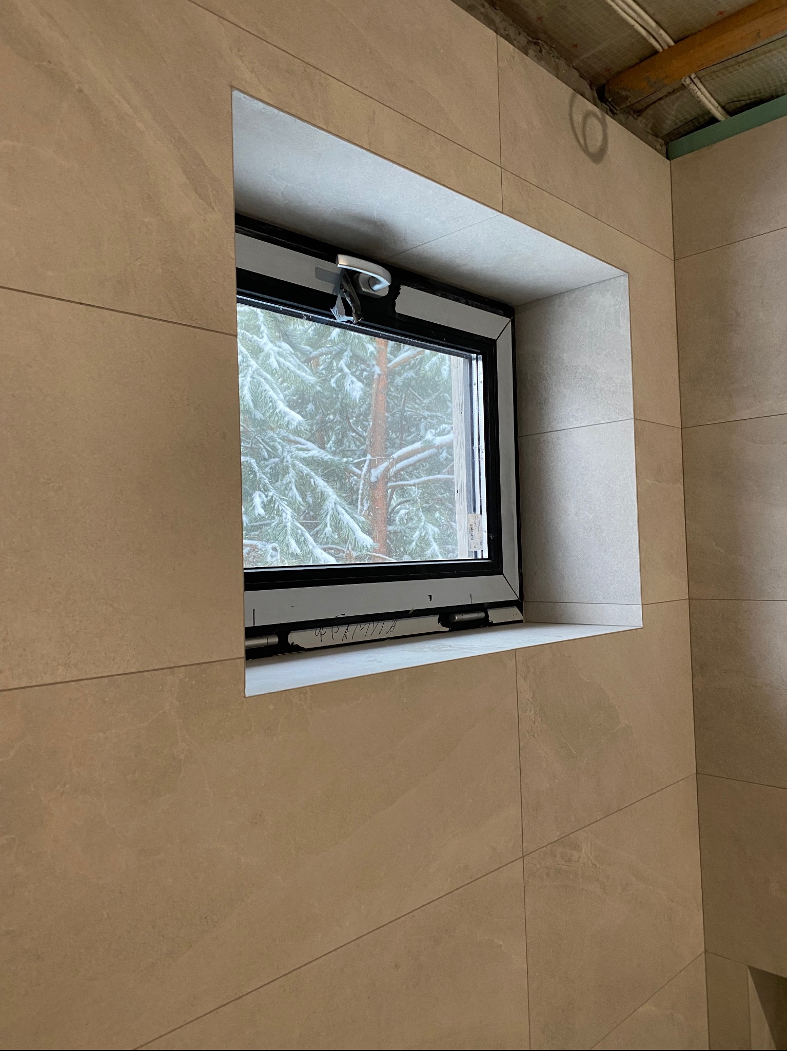 Jasne, beżowe płytki ceramiczne na ścianie łazienki, z oknem na zimowy krajobraz za szybą. Widoczne detale montażu okna i fugi płytek.