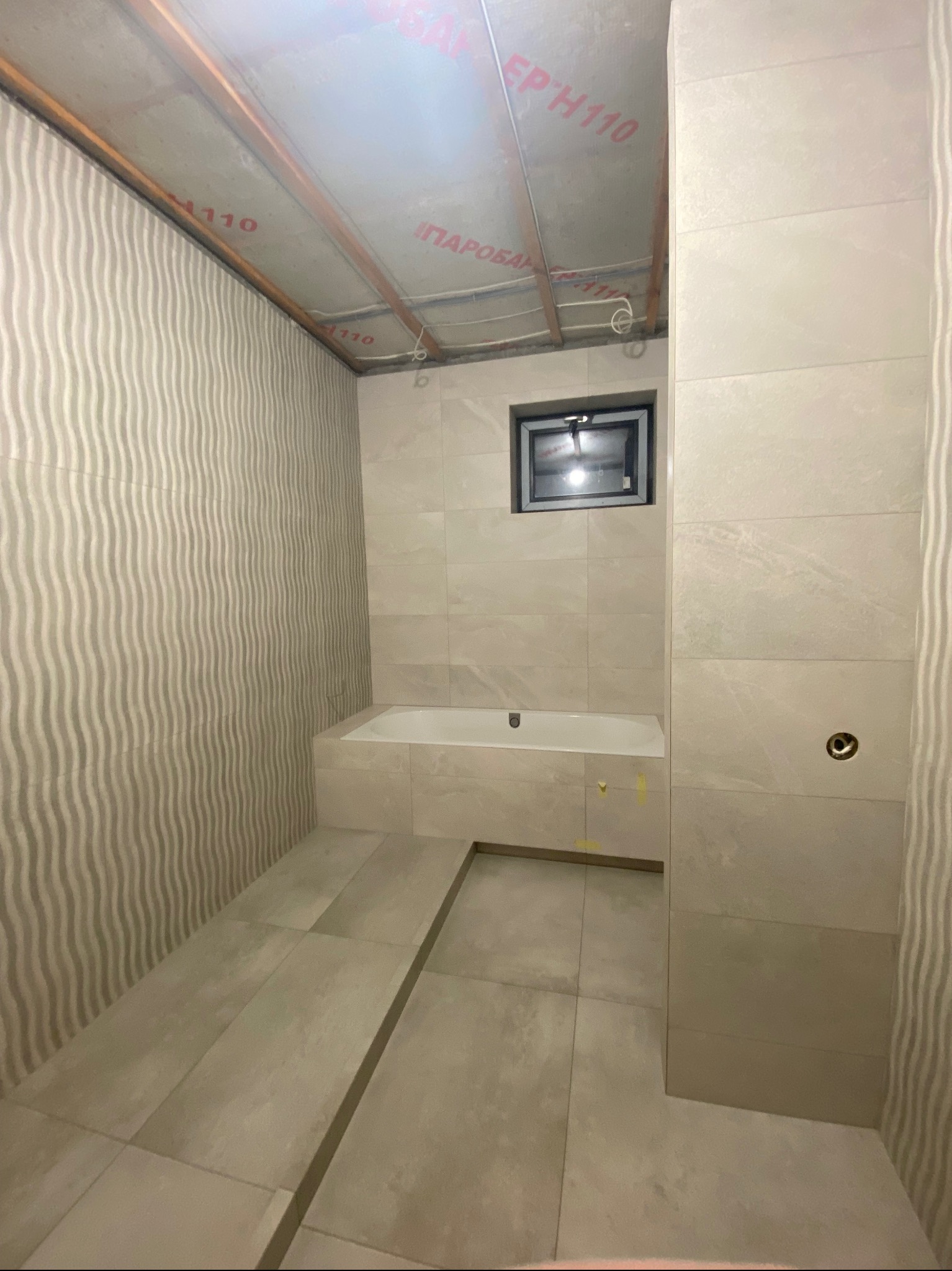 Wnętrze łazienki w trakcie prac wykończeniowych; widoczne płytki na ścianach z subtelnym wzorem falistym, zabudowana wanna, małe okno i odsłonięty sufit z instalacjami.