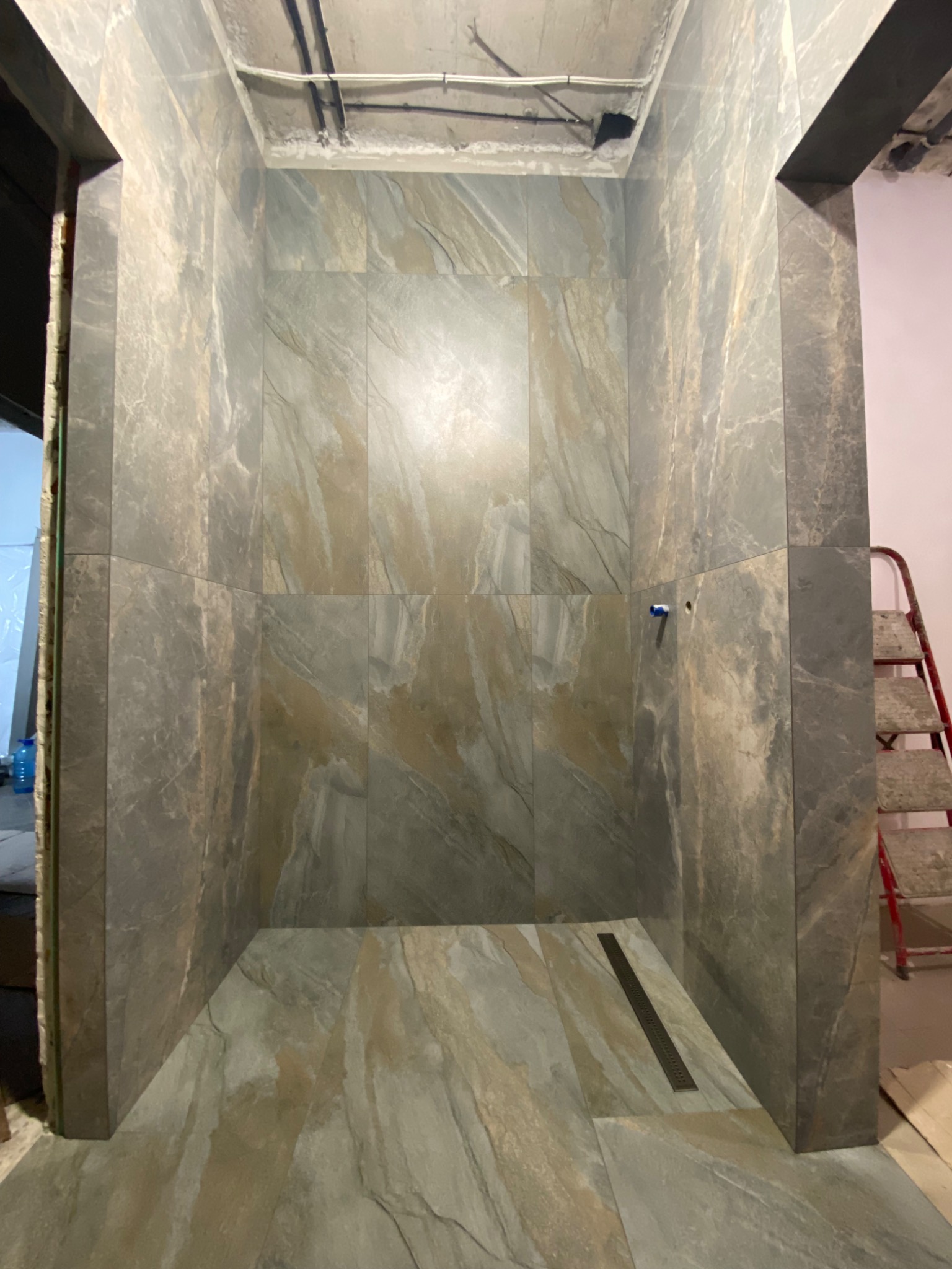 Wykończony prysznic typu walk-in z szaro-beżowymi płytkami imitującymi kamień, widoczny odpływ liniowy w podłodze, surowy sufit z instalacjami, drabina malarska w tle.