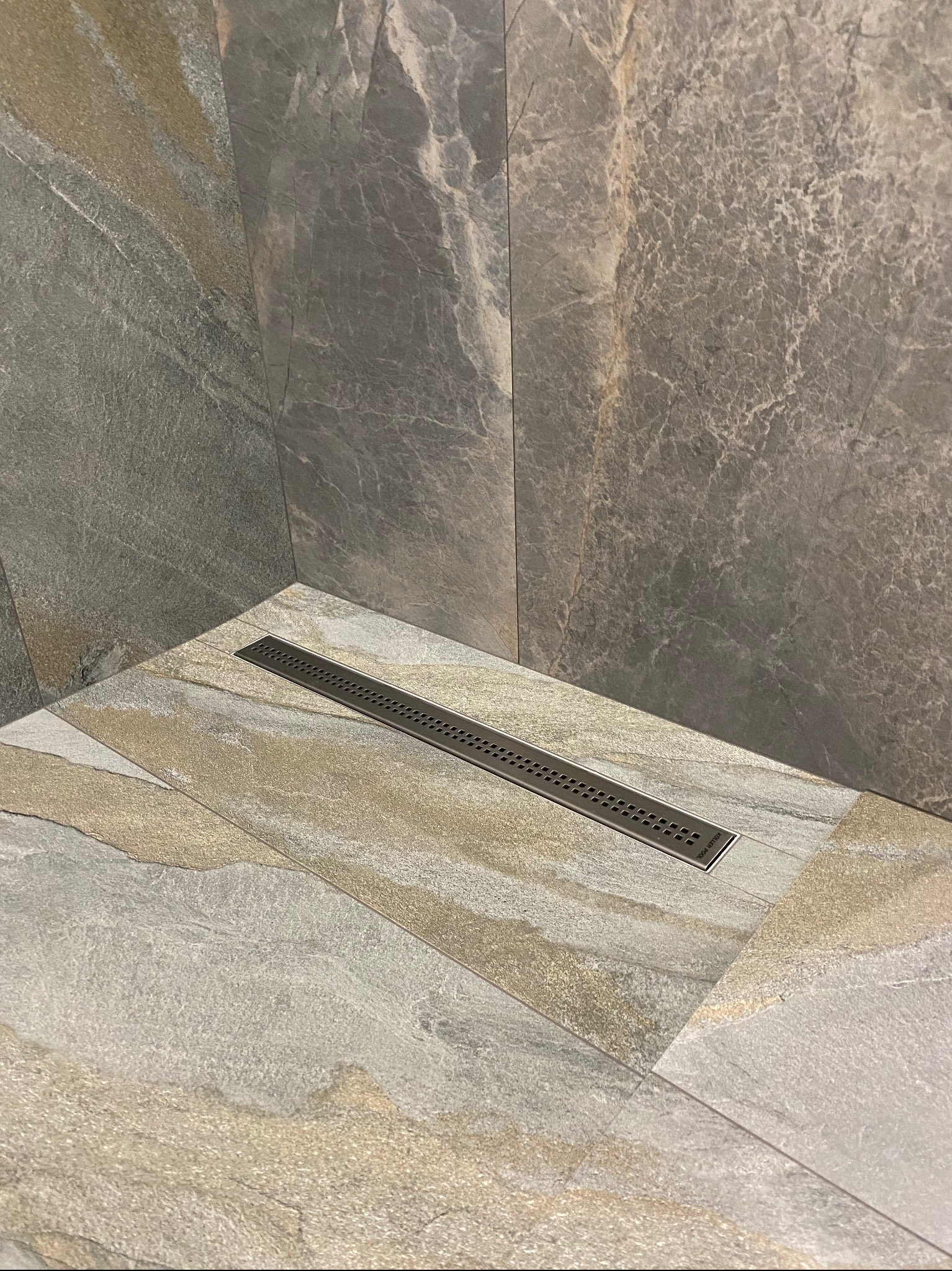 Nowoczesny odpływ liniowy w kabinie prysznicowej wyłożonej płytkami imitującymi kamień w odcieniach szarości i beżu.