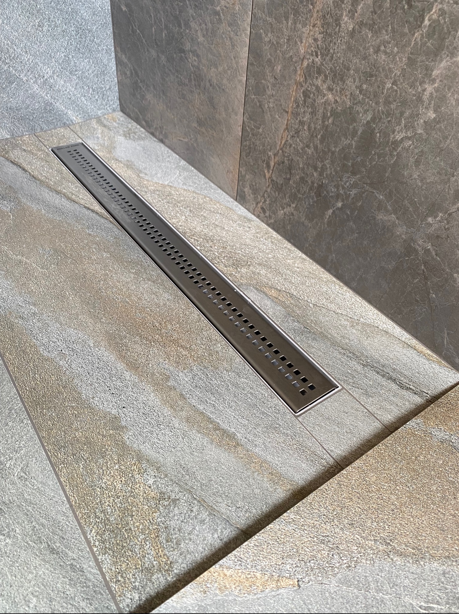 Nowoczesny odpływ liniowy w kabinie prysznicowej wyłożonej płytkami imitującymi kamień w odcieniach szarości i beżu.