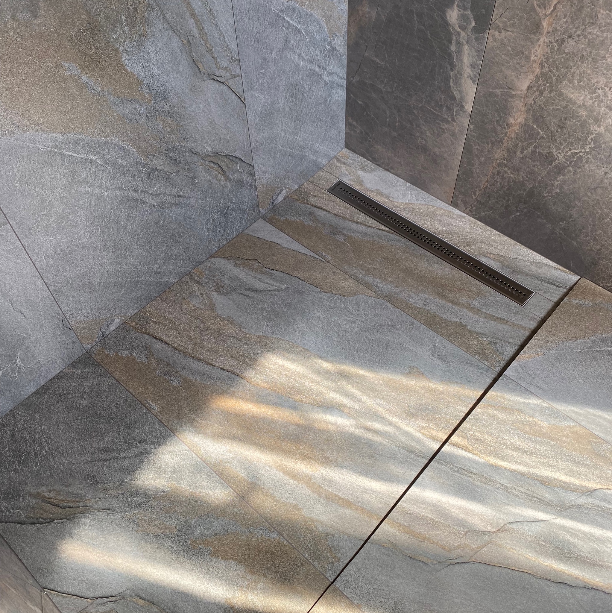 Szare i beżowe płytki imitujące kamień w kabinie prysznicowej z odpływem liniowym, oświetlone promieniami słońca.