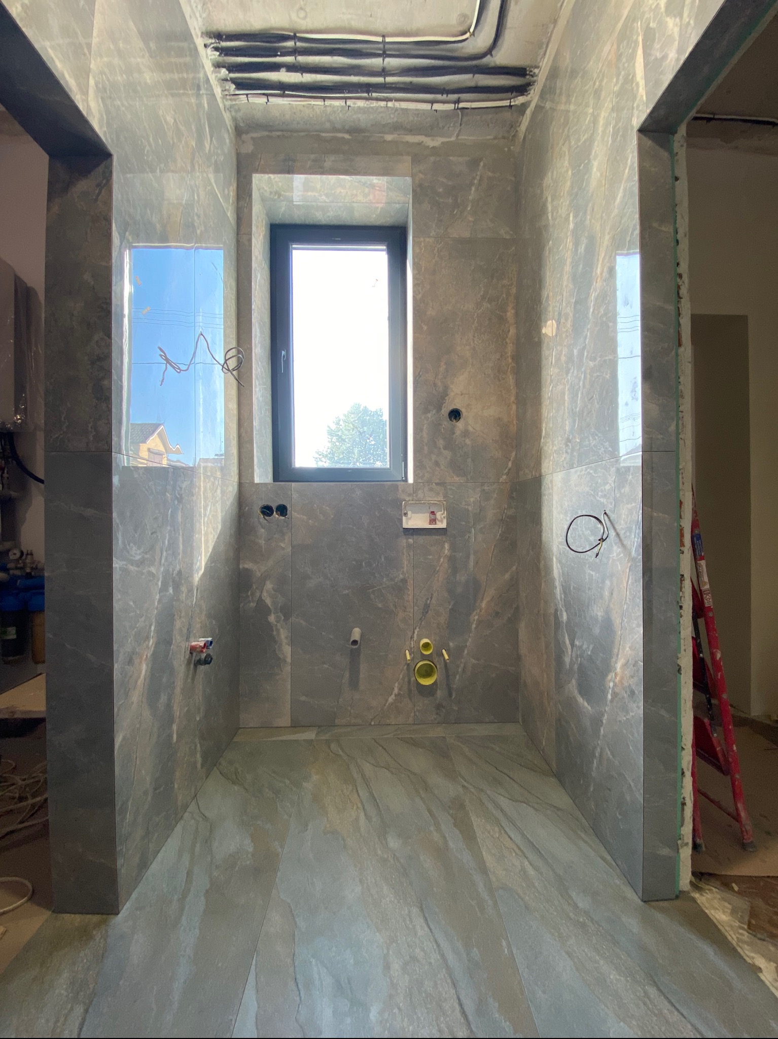 Wnętrze łazienki w trakcie wykańczania, szare płytki imitujące marmur na ścianach i podłodze, widoczne instalacje, okno z widokiem na zewnątrz, drabina oparta o ścianę.