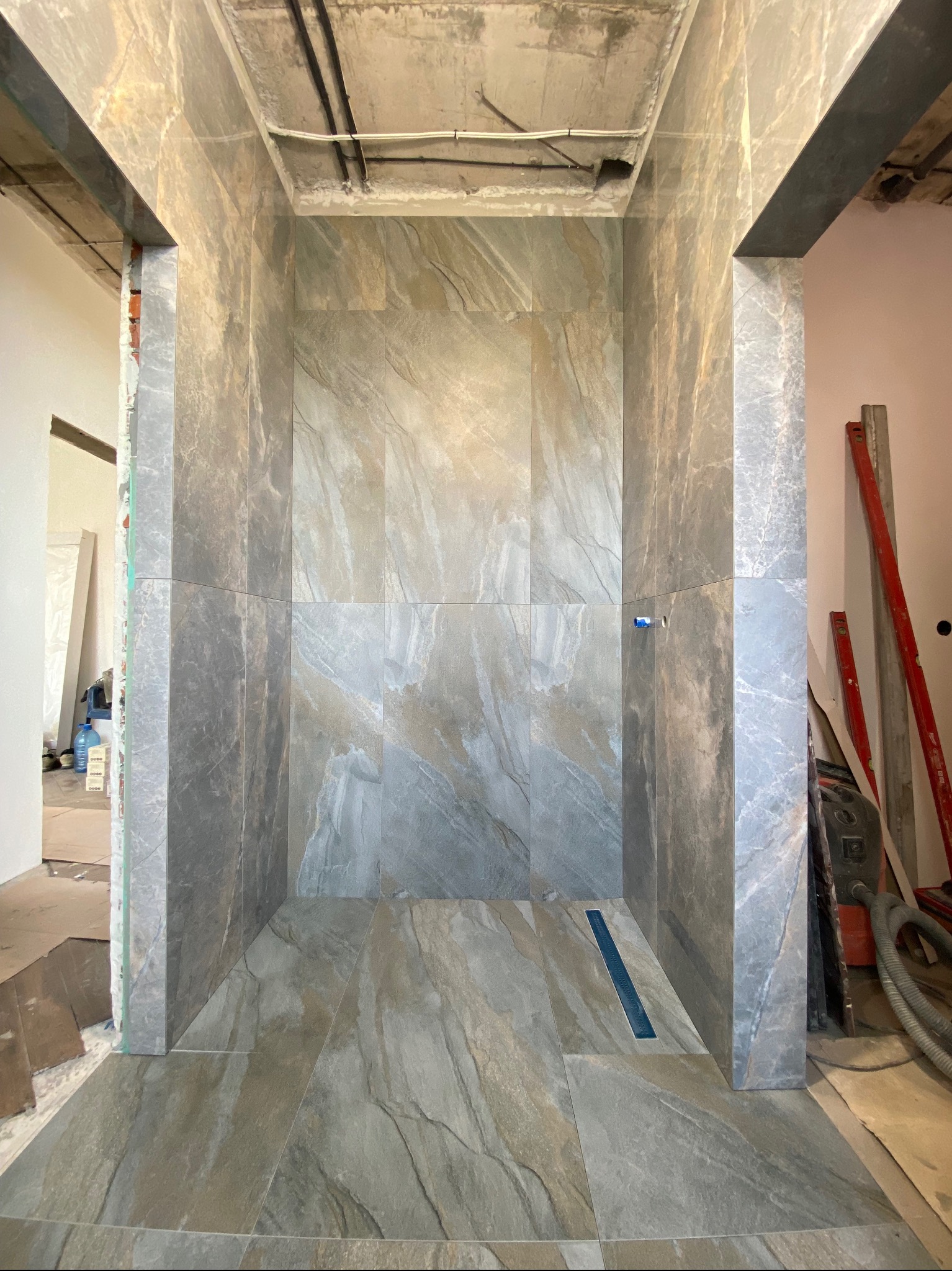 Wnętrze kabiny prysznicowej w trakcie wykańczania, z widocznymi płytkami imitującymi kamień w odcieniach szarości i beżu, niezakryty sufit z instalacjami, oraz odpływ liniowy w podłodze.