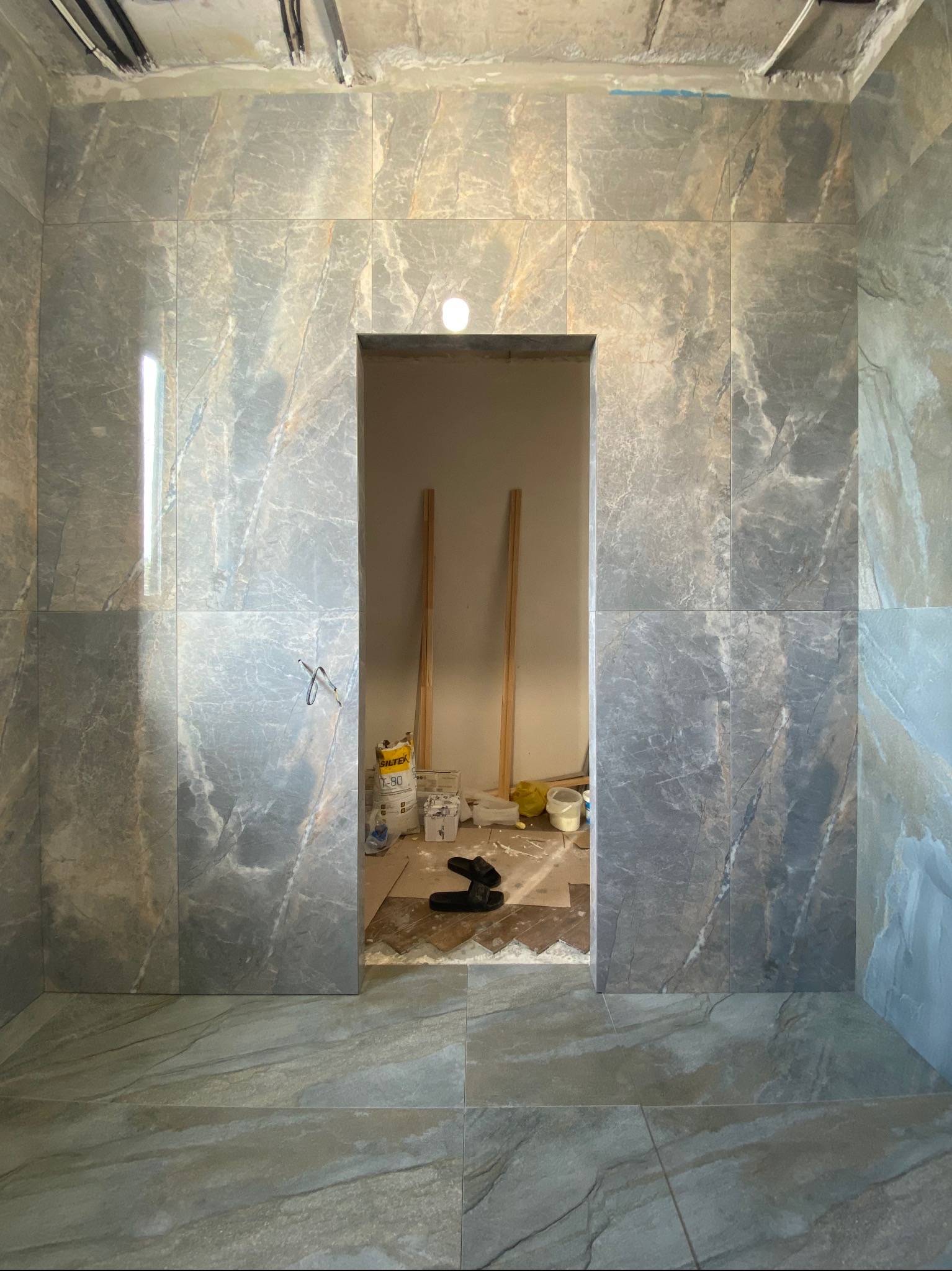 Łazienka w trakcie remontu, szare płytki imitujące marmur na ścianach i podłodze, widoczne narzędzia i materiały budowlane w przejściu do kolejnego pomieszczenia, surowy sufit z instalacjami.