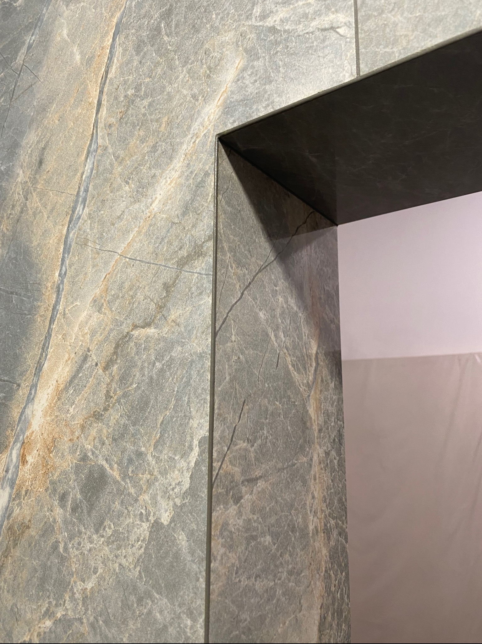 Fragment ściany wyłożonej szarą marmurkową płytką w łazience, z widocznym narożnikiem obłożonym identyczną płytką, tworzącym obramowanie przejścia.