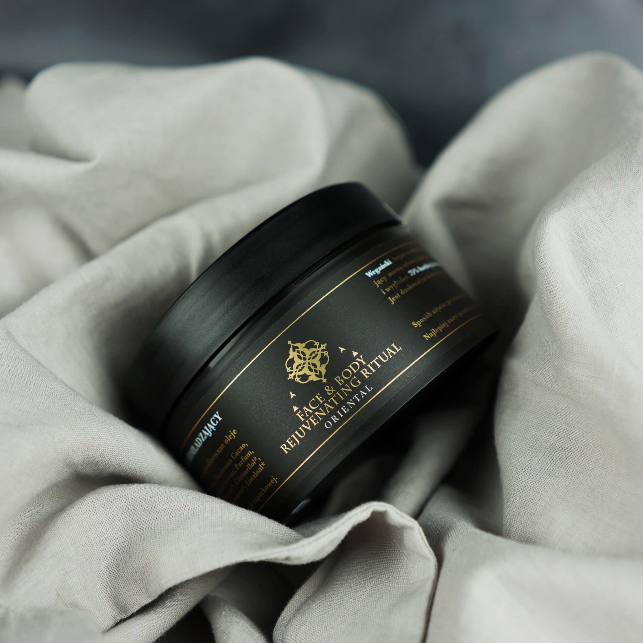 Czarny słoiczek z kremem do twarzy i ciała z orientalnym motywem, umieszczony na delikatnej, lnianej tkaninie w odcieniach szarości, ukazujący detale etykiety z informacjami o produkcie.