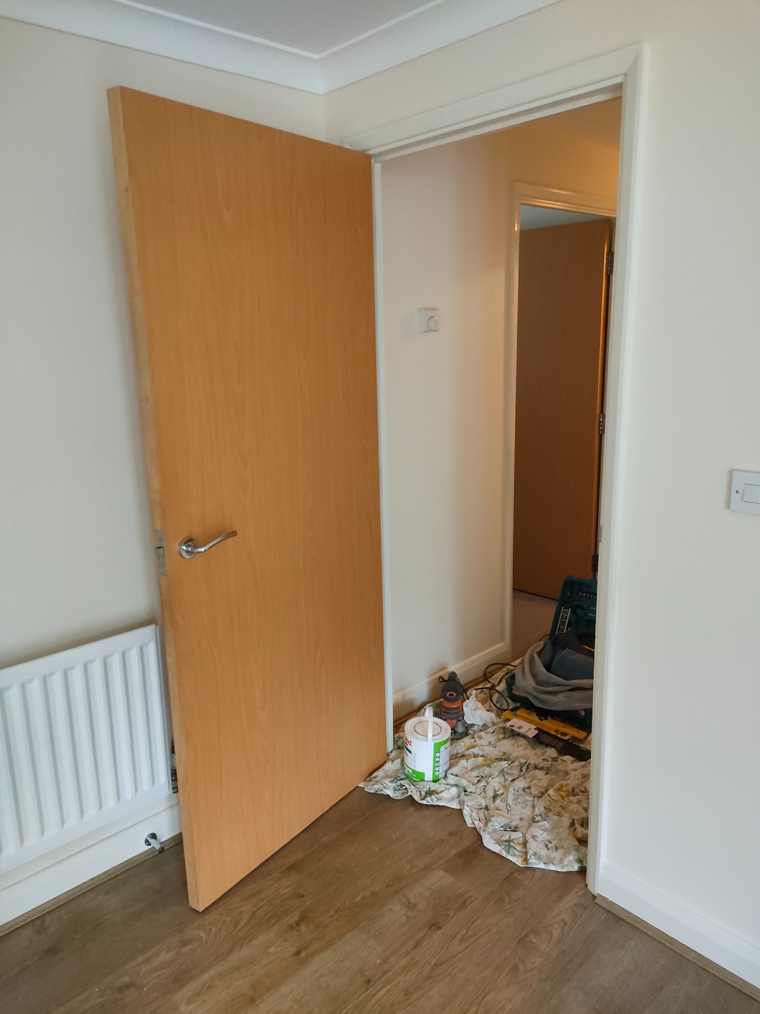 Montaż drzwi wewnętrznych z drewnianą okleiną, widoczny fragment grzejnika i resztki prac remontowych w przejściu do drugiego pomieszczenia.