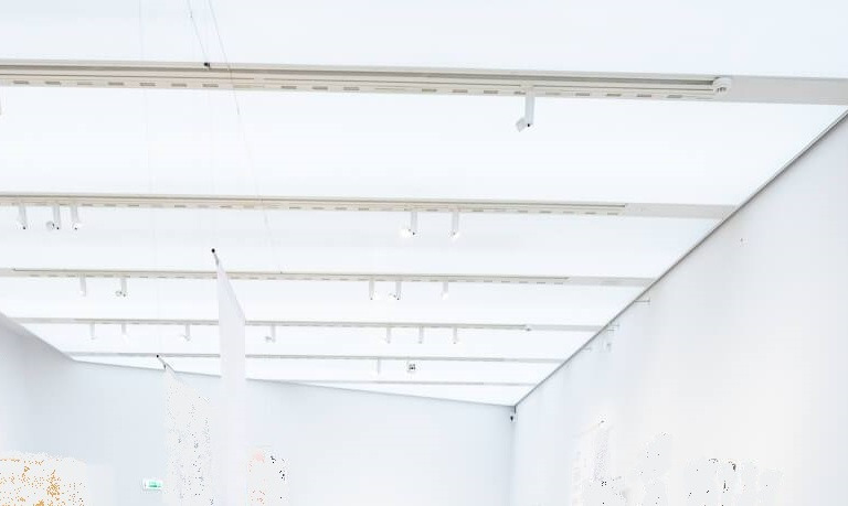 Sufit galerii z zamontowanym systemem szyn oświetleniowych i reflektorami punktowymi, widok z dołu, jasne wnętrze.