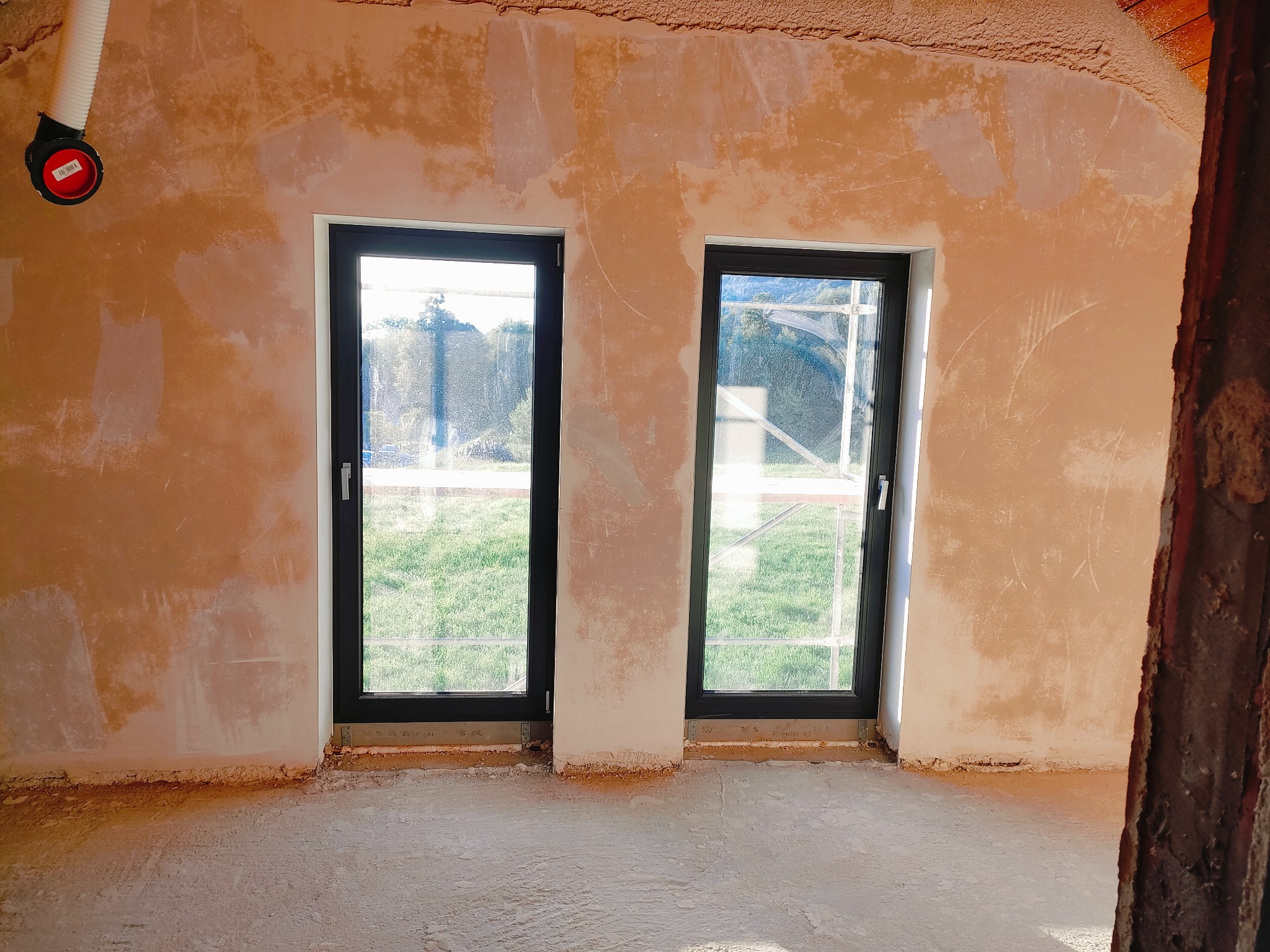 Wnętrze w trakcie prac wykończeniowych: surowe ściany po nałożeniu tynku, widoczne dwa okna z czarnymi ramami, podłoga wylewka, na ścianie zawieszony przewód wentylacyjny.