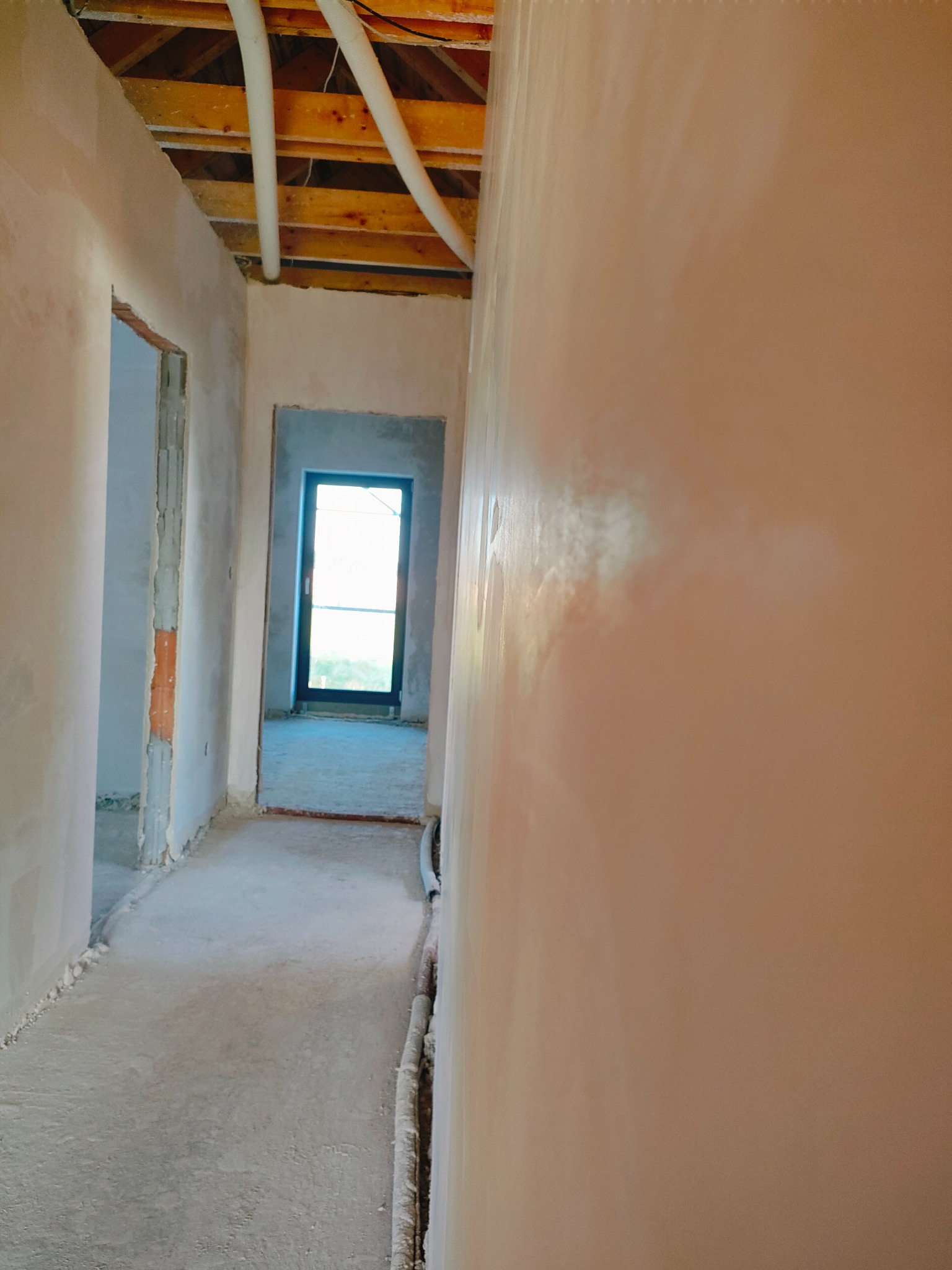 Korytarz w trakcie prac wykończeniowych; świeżo nałożone jasnoróżowe gładzie szpachlowe na ścianach, surowy beton na podłodze, widoczne rury pod sufitem i ościeżnice drzwiowe w stanie surowym.