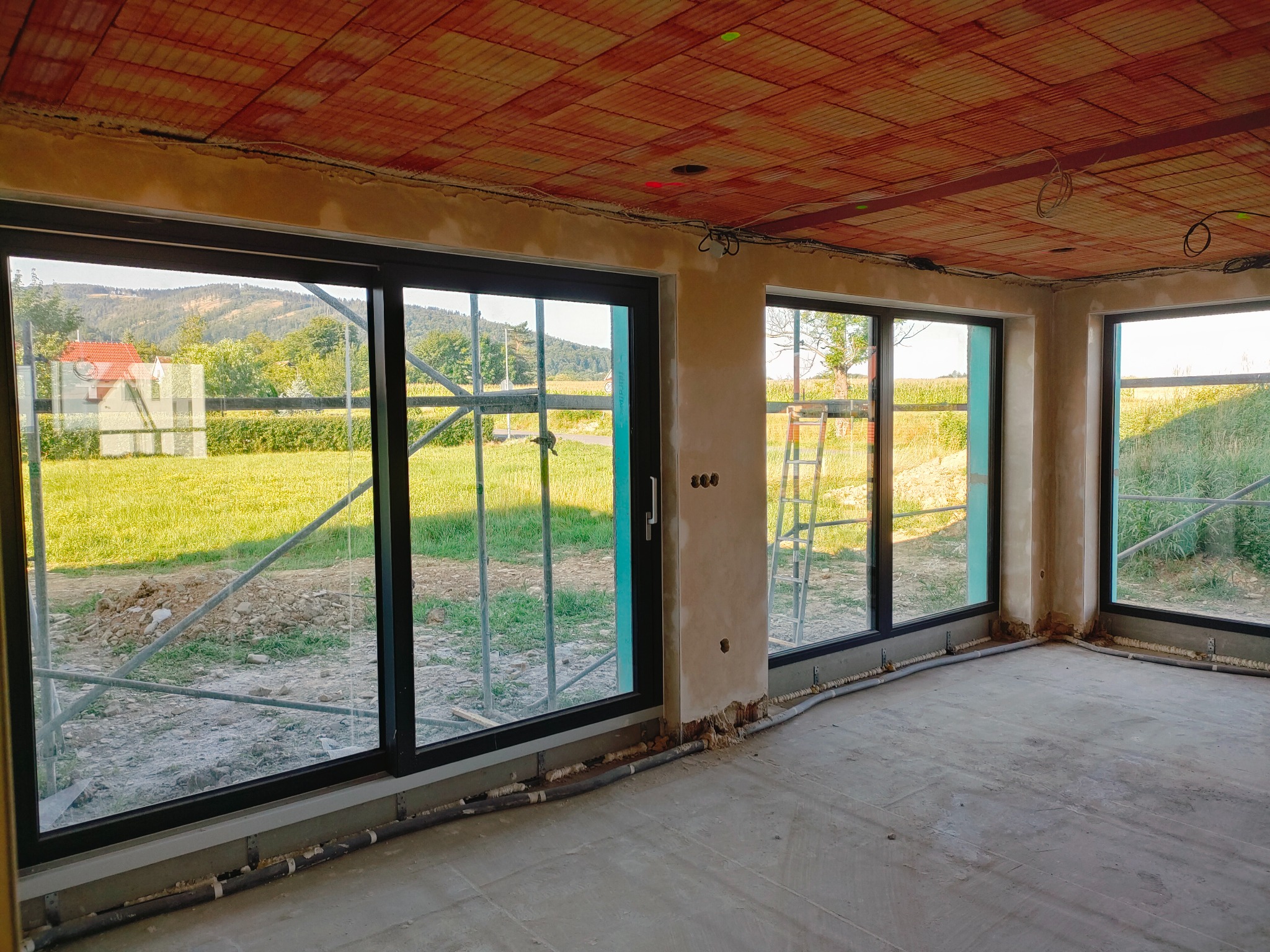 Surowe wnętrze domu w trakcie prac wykończeniowych, widoczne okna z czarnymi ramami, ściany częściowo pokryte tynkiem, instalacje pod sufitem i na podłodze, oraz ceglany strop.