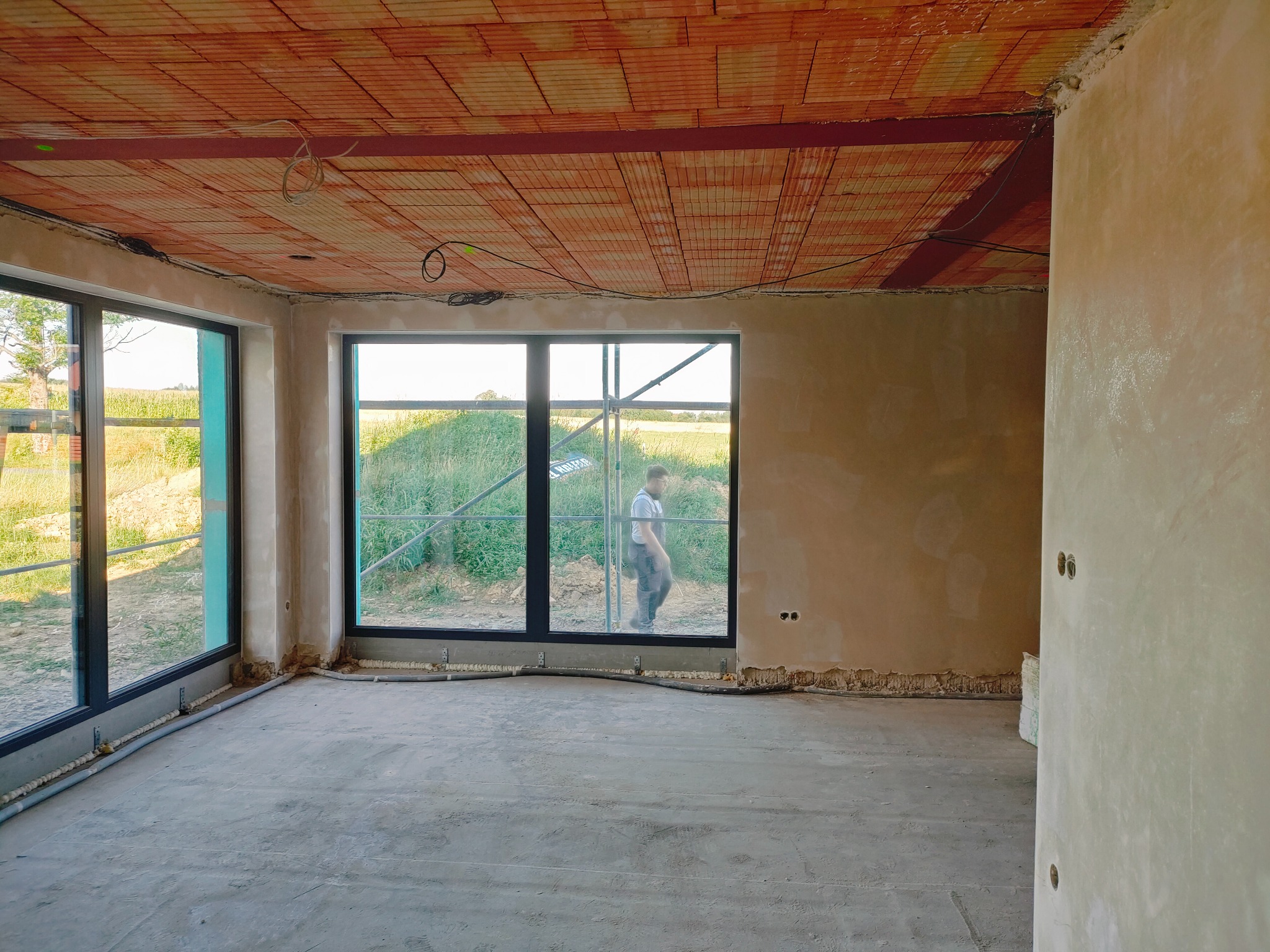 Surowy stan wnętrza z widocznymi ceglanymi stropami, wstępnie otynkowanymi ścianami, dużymi oknami z widokiem na zewnątrz i pracownikiem na rusztowaniu za oknem.