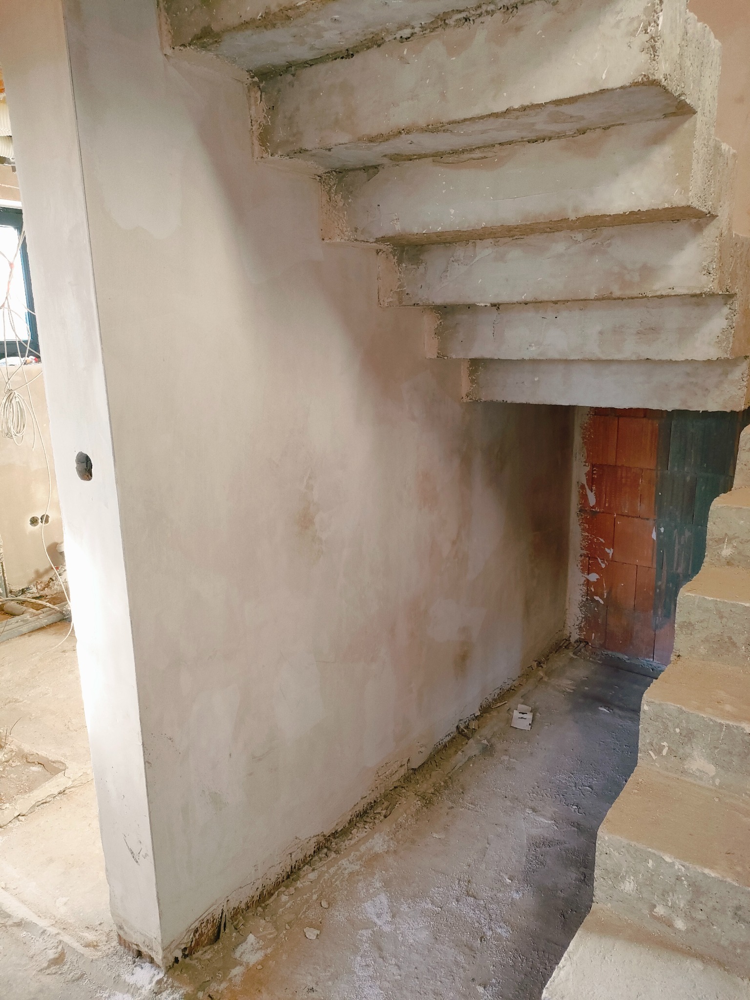 Wnętrze w trakcie prac wykończeniowych: ściana po nałożeniu gładzi gipsowej, surowe betonowe schody z widocznymi stopniami, fragment ściany z cegły w głębi.