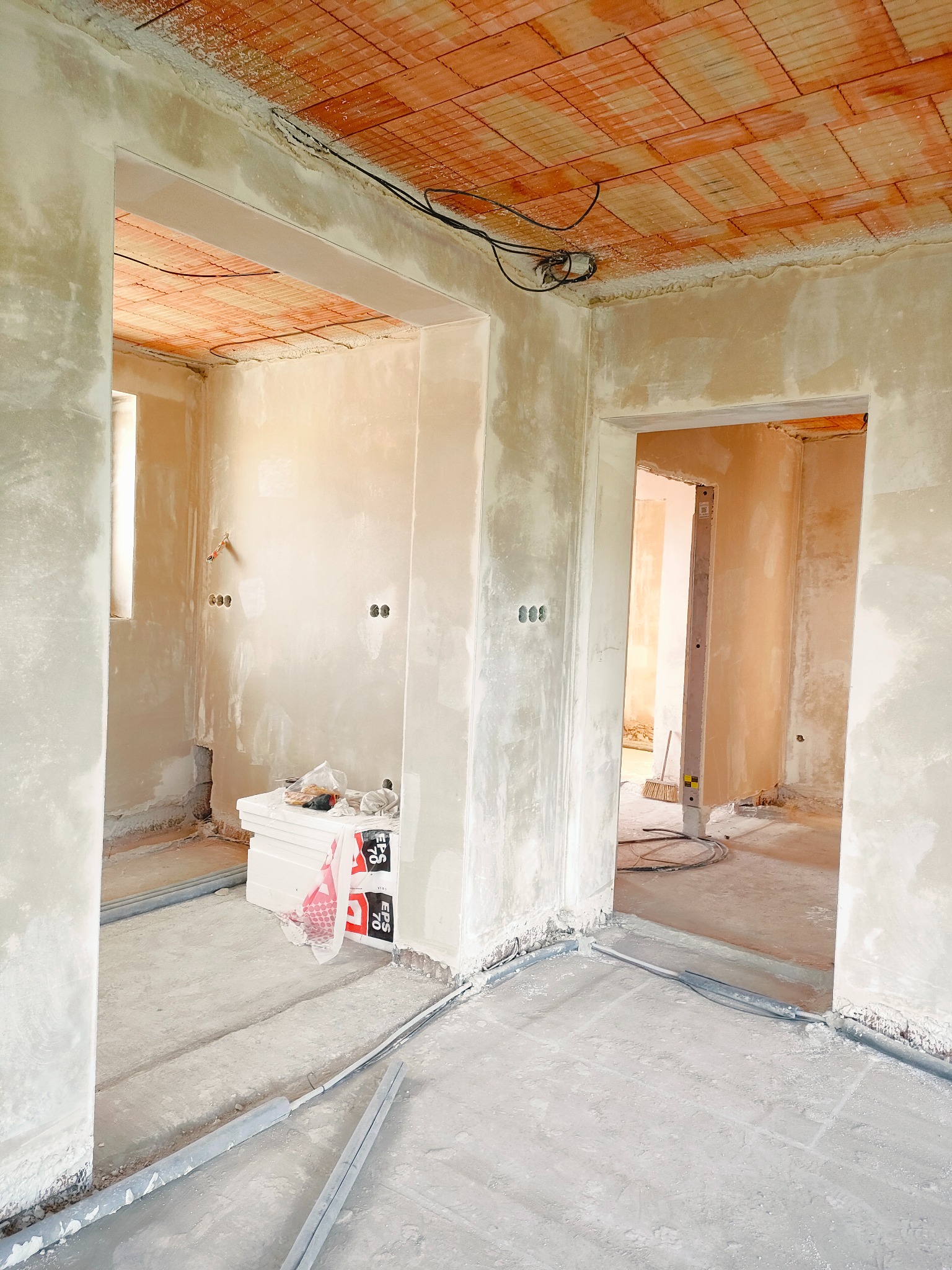 Surowe wnętrze w trakcie prac wykończeniowych: ściany pokryte świeżym tynkiem, widoczne instalacje elektryczne, ceglany strop i materiały izolacyjne na podłodze, zarys dwóch przejść do kolejnych...