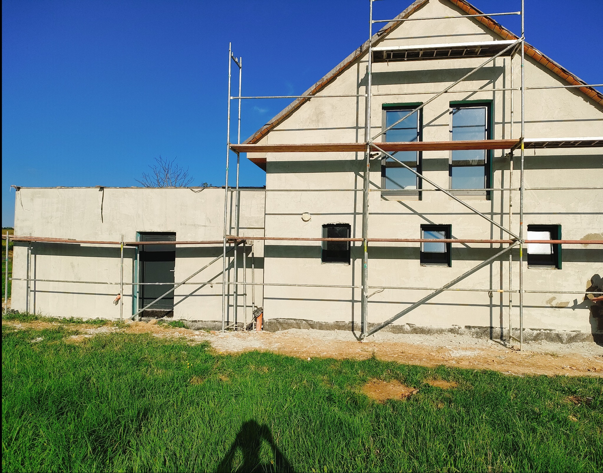 Dom w trakcie nakładania tynków zewnętrznych, widoczne rusztowania, świeżo otynkowana elewacja z oknami i drzwiami, trawa w dolnej części kadru, błękitne niebo.