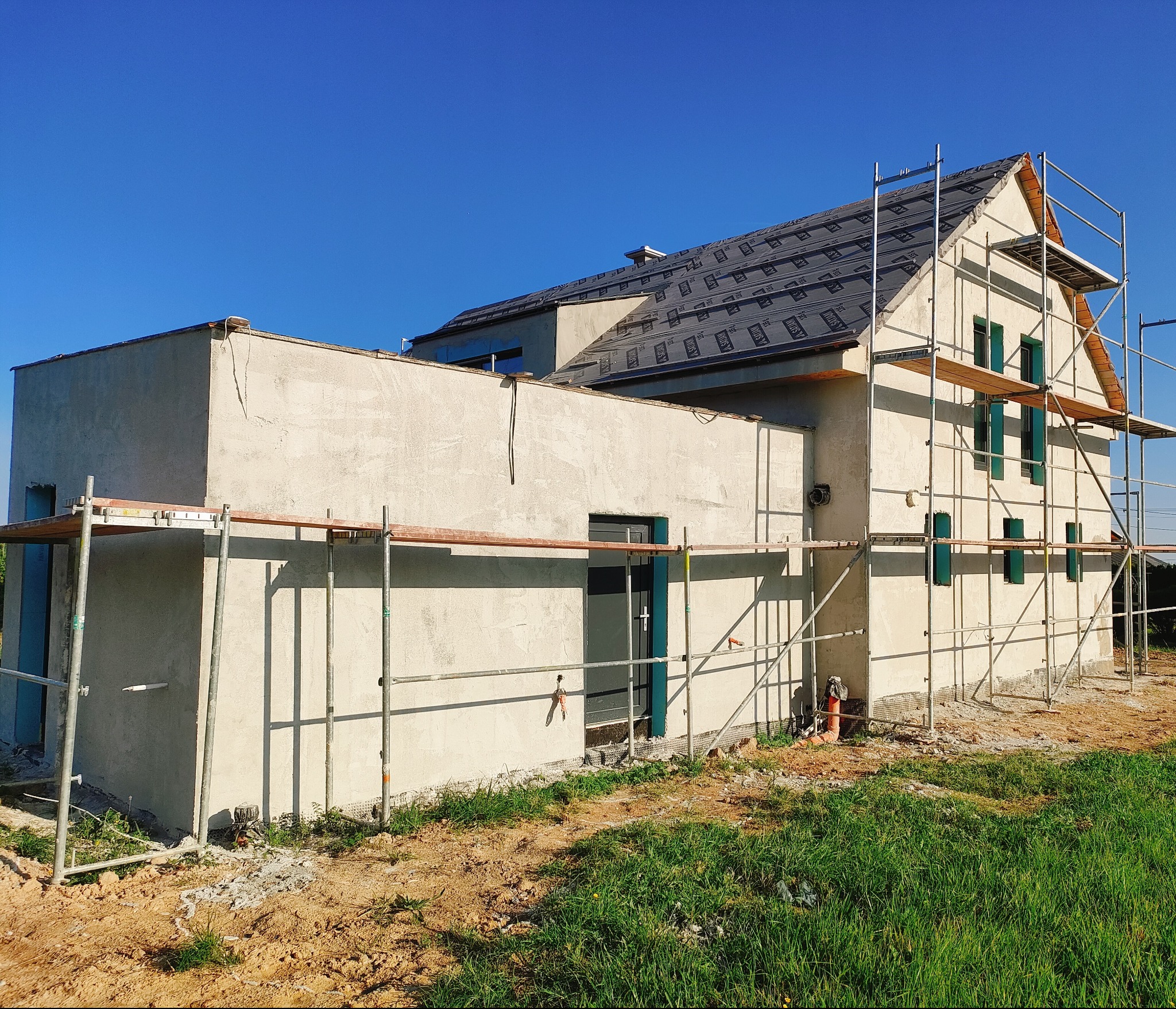 Dwukondygnacyjny dom w trakcie nakładania tynku maszynowego, widoczne rusztowania i wstępnie otynkowana elewacja w jasnym odcieniu, dach pokryty membraną dachową, błękitne niebo w tle.