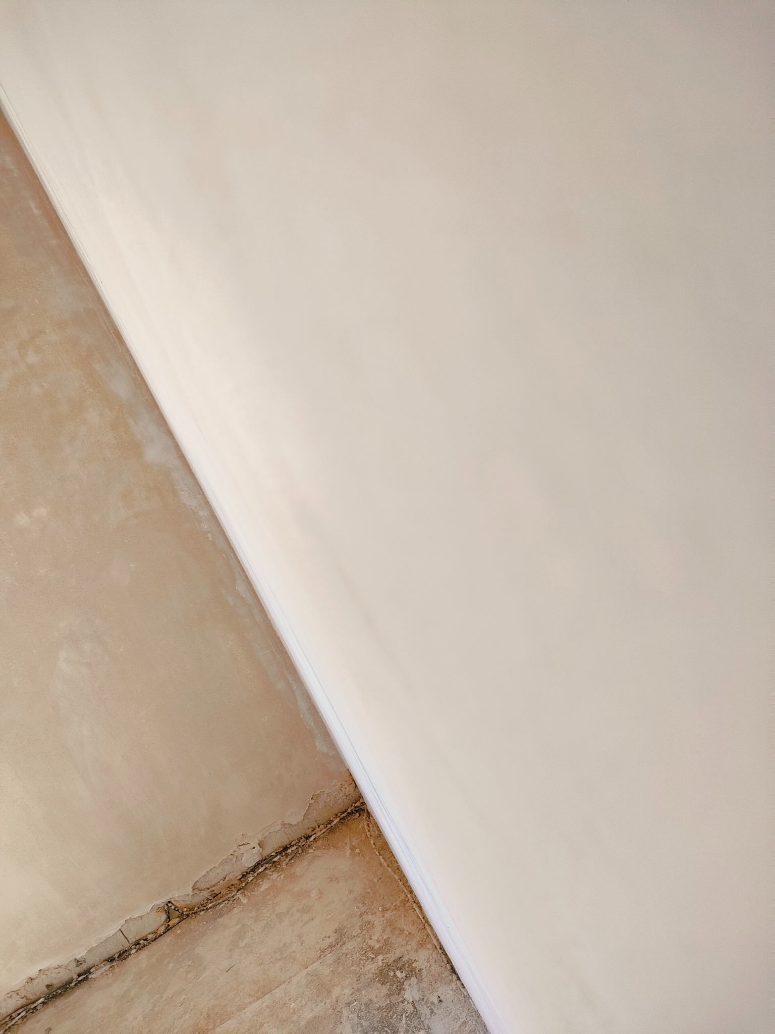 Fragment ściany po tynkowaniu maszynowym, widoczna gładka, jasna powierzchnia tynku oraz surowa podłoga z widocznymi pęknięciami i pozostałościami po pracach budowlanych.