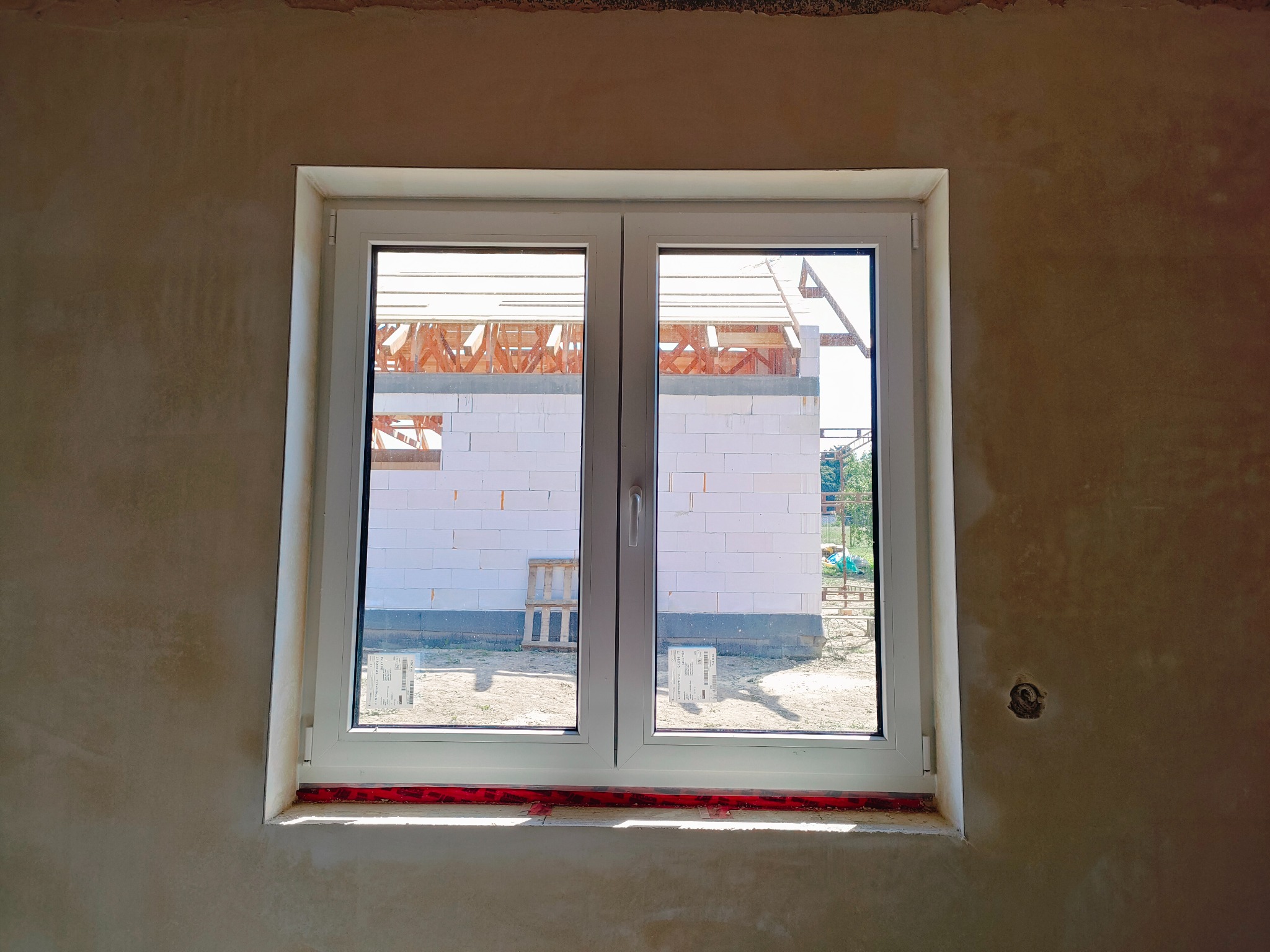 Surowa ściana z nałożonym gruntem pod tynk, z widocznym oknem, przez które widać budowę domu w stanie surowym otwartym z białego bloczka.