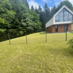 Nowoczesny dom z dużym oknem na wzgórzu, otoczony świeżo skoszoną trawą i gęstym lasem, ogrodzony siatką.