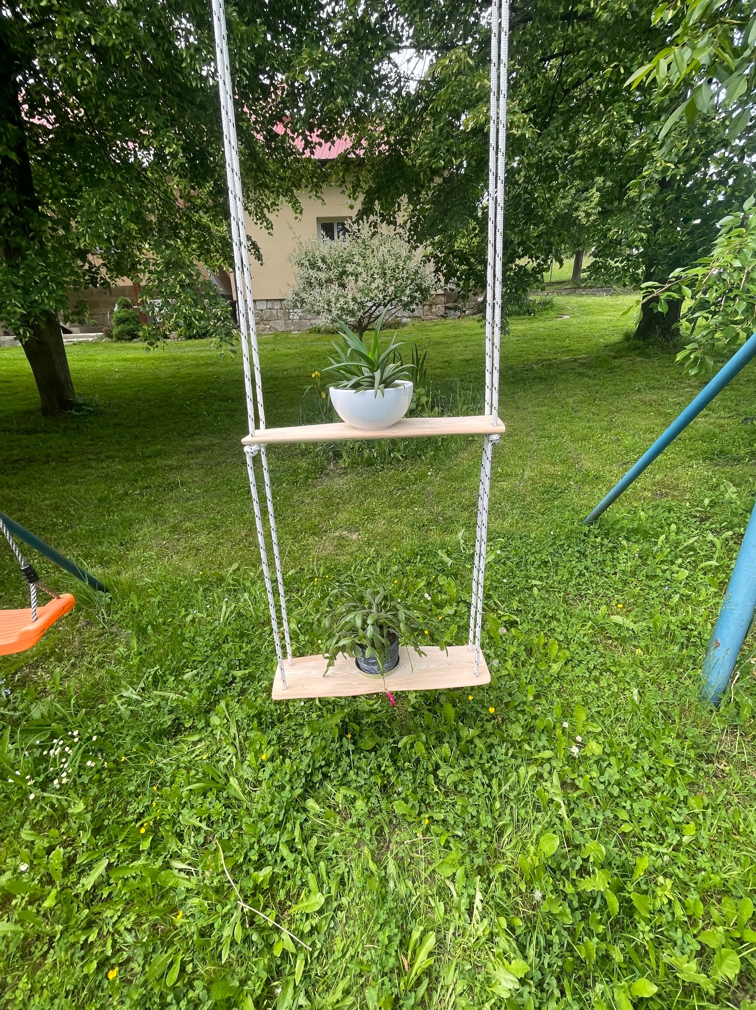 Dwu-poziomowa huśtawka z jasnego drewna zawieszona na grubych, białych linach, służąca jako kwietnik na tle zielonego trawnika z domem w tle.