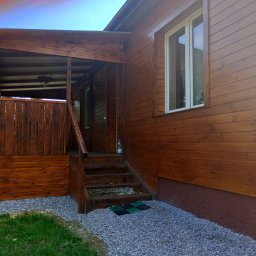 Łukasz Stąpór. TURBOBUD - Drewniany dom z zadaszonym wejściem i schodami, świeżo pomalowany na brązowo, otoczony szarym żwirem.