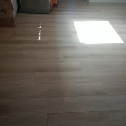 Łukasz Stąpór. TURBOBUD - Jasne panele podłogowe ułożone w kuchni, z widocznym fragmentem szafki i torbą na zakupy w rogu, oświetlone silnym, naturalnym światłem słonecznym.