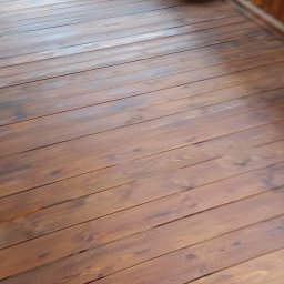 Łukasz Stąpór. TURBOBUD - Drewniana podłoga tarasu z widocznymi słojami i brązową bejcą, świeżo po renowacji, z fragmentem balustrady w tle.