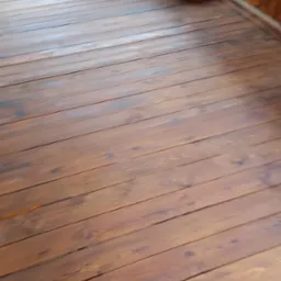 Drewniana podłoga tarasu z widocznymi słojami i brązową bejcą, świeżo po renowacji, z fragmentem balustrady w tle.