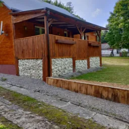 Drewniany dom z tarasem, podmurówka z gabionów wypełnionych białymi kamieniami, widok z zewnątrz na tle zieleni.