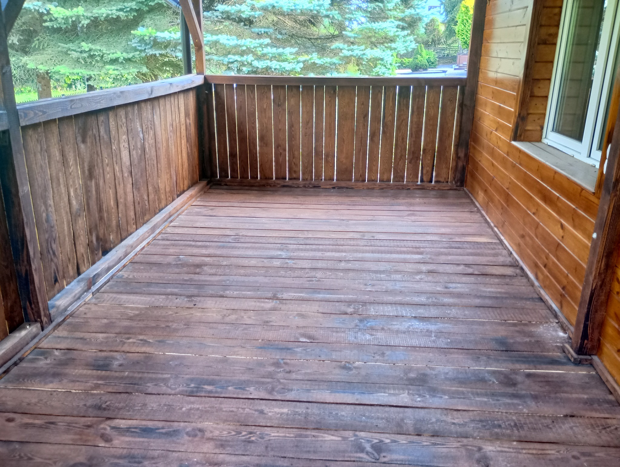 Drewniany taras z balustradą, widok na zieleń, świeżo po impregnacji, deski podłogowe z widocznym usłojeniem.