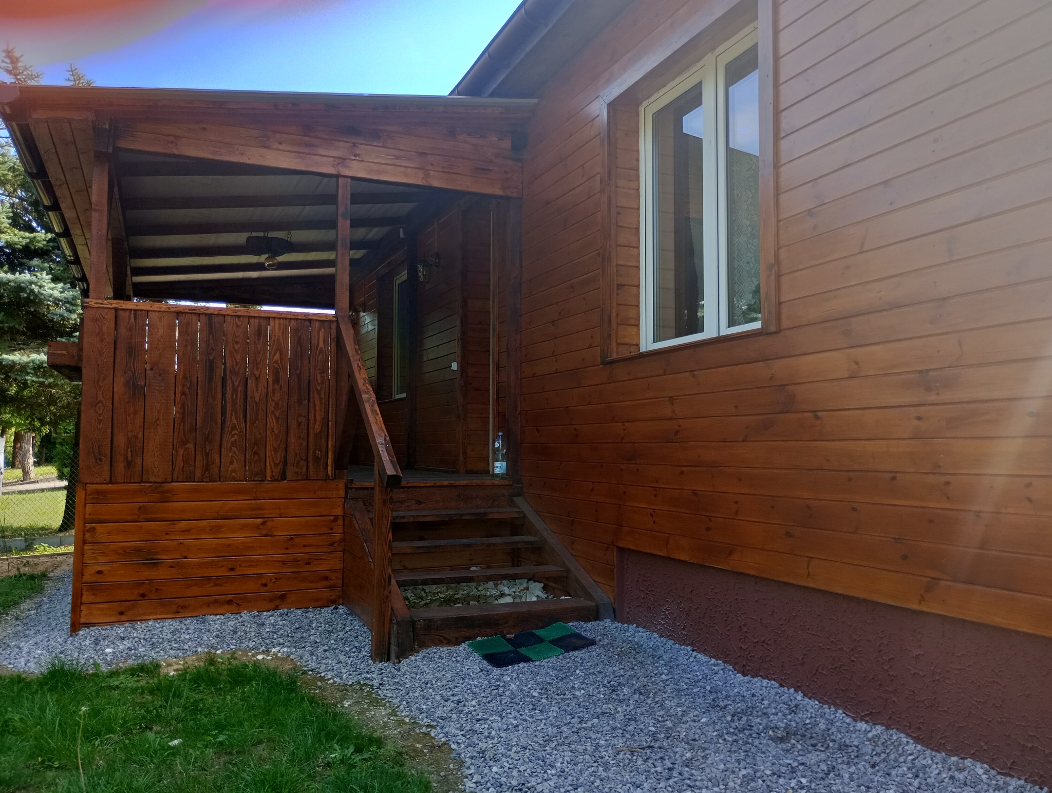 Drewniany dom z zadaszonym wejściem i schodami, świeżo pomalowany na brązowo, otoczony szarym żwirem.